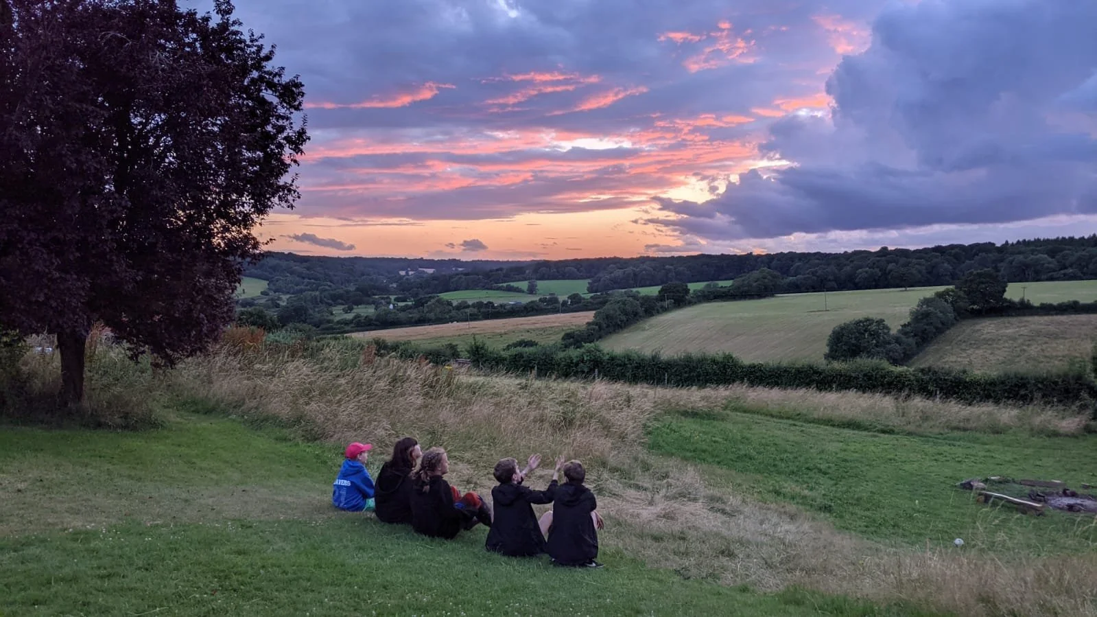 A group of five people sitting on a grassy hill, watching a sunset over rolling green fields and distant trees, with a partly cloudy sky illuminated in pink and purple hues.