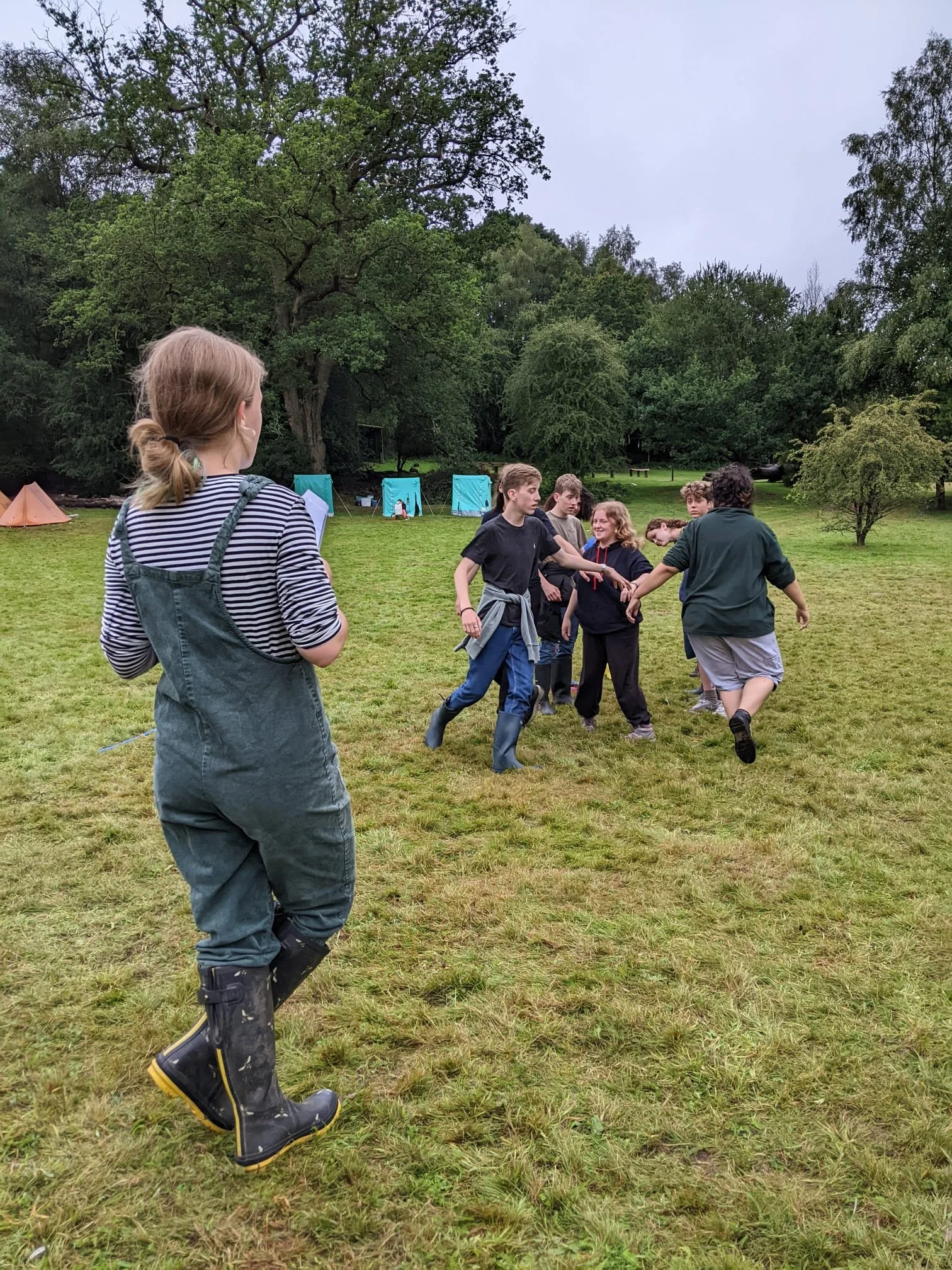 A group of children and teenagers playing and holding hands on a grassy field in a park with trees and tents in the background.