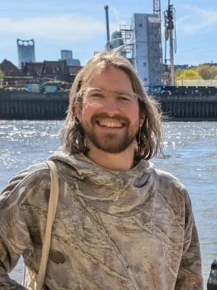 A smiling man with long hair and a beard standing outdoors near a body of water, with industrial buildings and factories in the background.