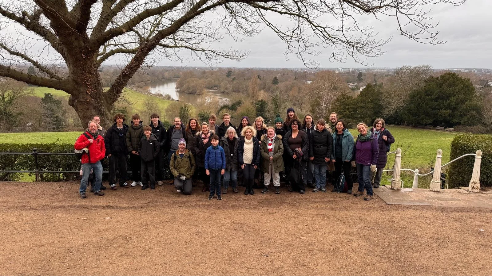 Group of people gathered outdoors in front of a scenic landscape with trees, a river, and cloudy sky.