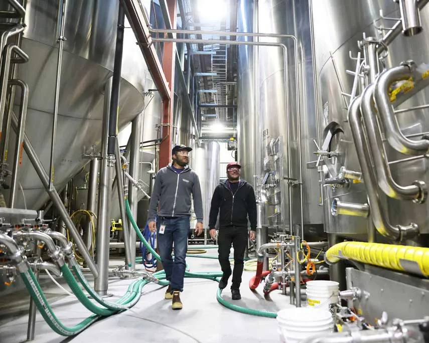 Two men walking through a large industrial brewery or food processing facility with stainless steel tanks and pipes.