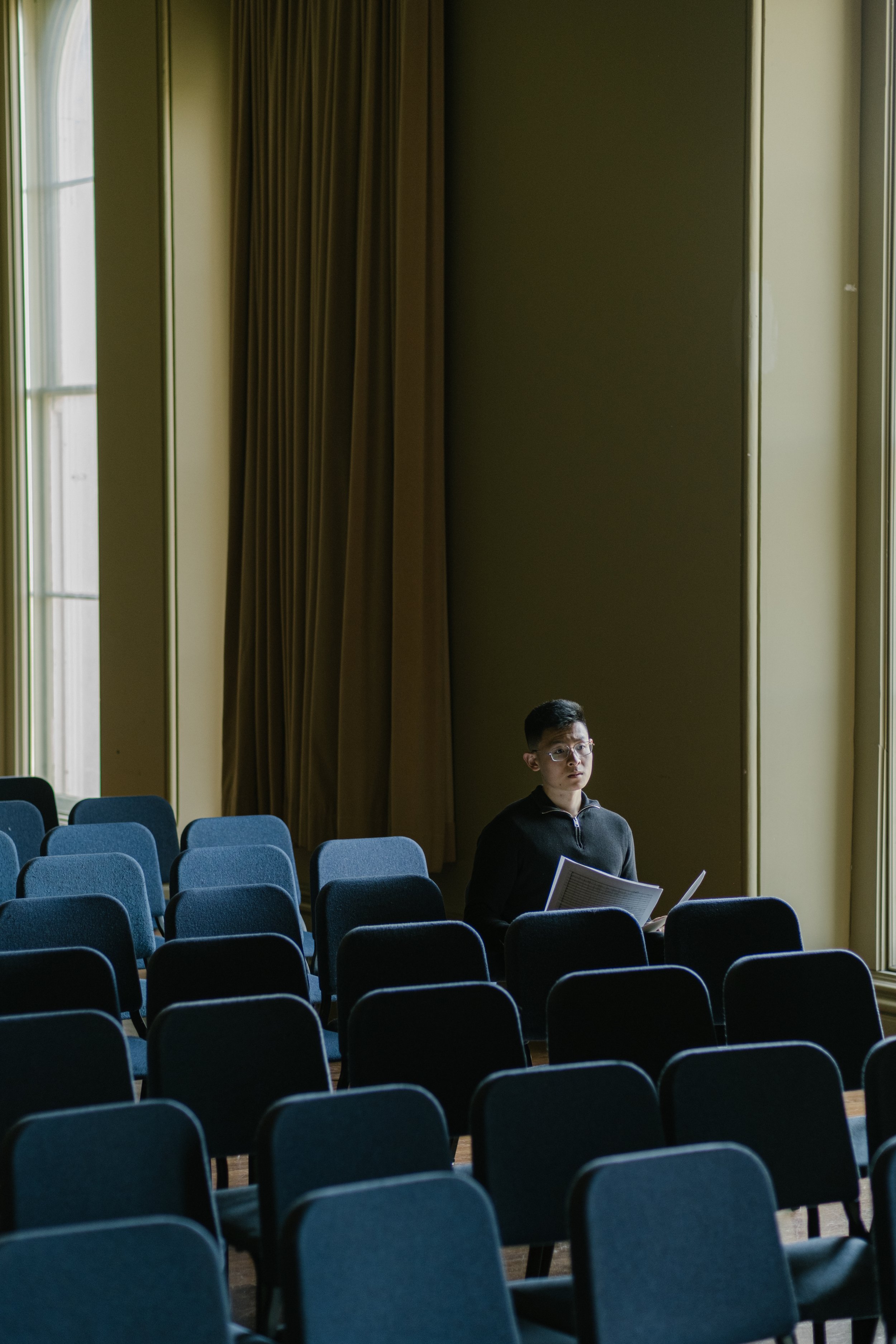 A young man with glasses sitting in a mostly empty auditorium or conference room, holding papers, with rows of blue chairs and large windows with curtains in the background.