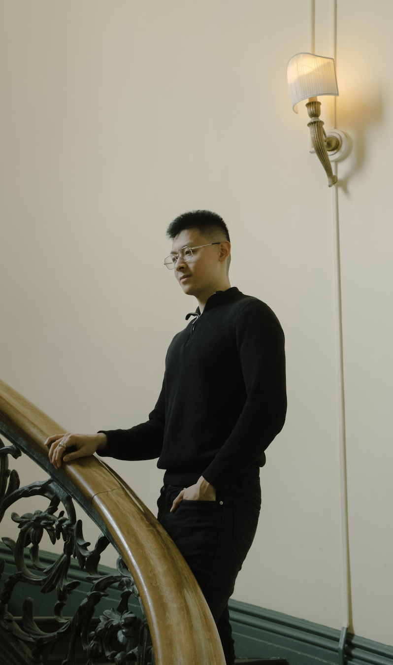 A young man with glasses and short black hair standing on a staircase, resting his hand on the wooden handrail, with a lit wall sconce above him, against a light-colored wall.