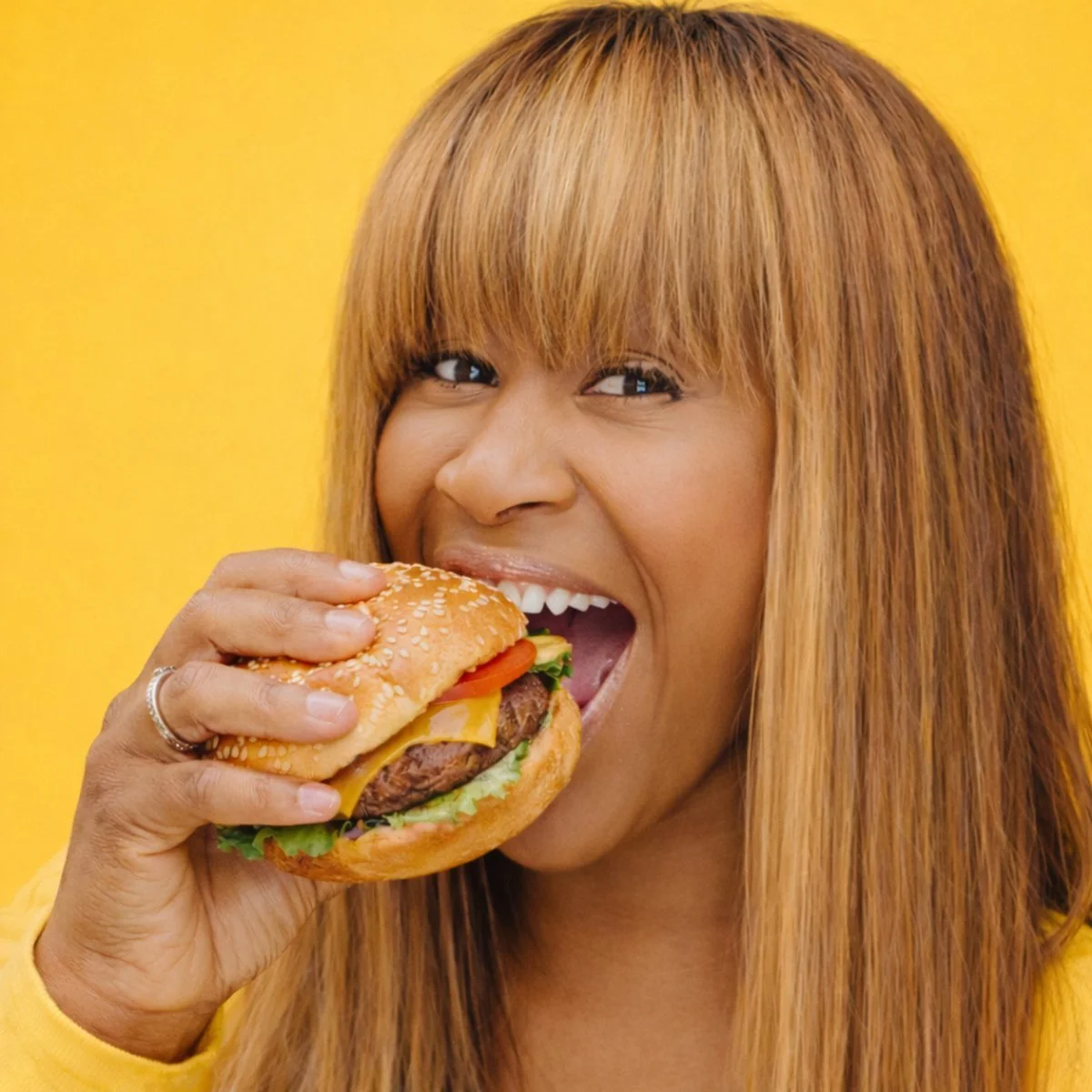 Woman with long hair biting into a cheeseburger with lettuce, tomato, cheese, and a sesame seed bun against a yellow background.