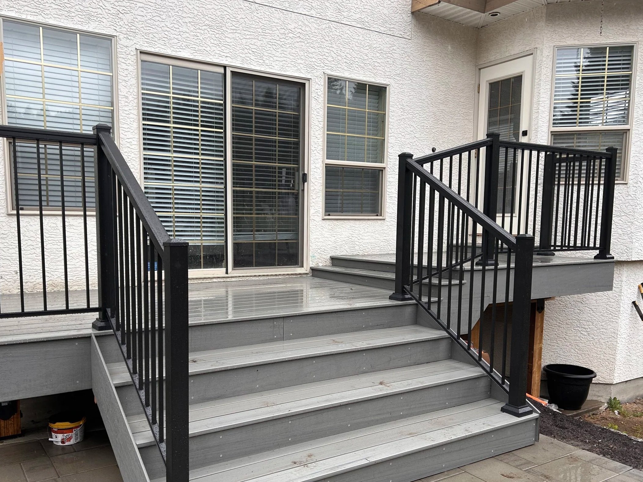 Backyard patio with gray wooden stairs and black metal railings leading to a house with white stucco exterior and large windows with white blinds.