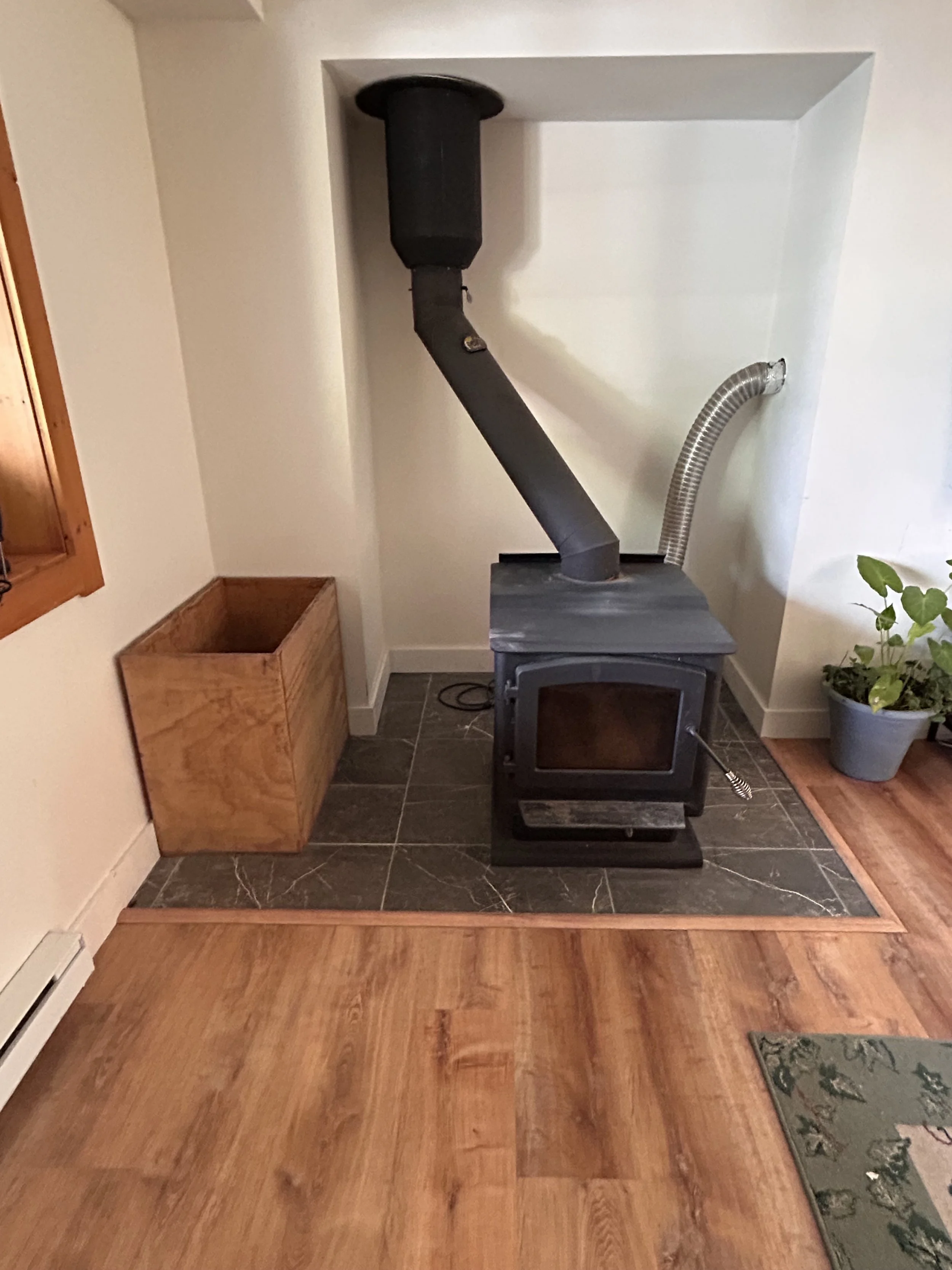 A black wood stove with a chimney pipe and flexible vent hose on a tiled hearth, next to a potted plant and a wooden storage box in a room with hardwood floors.