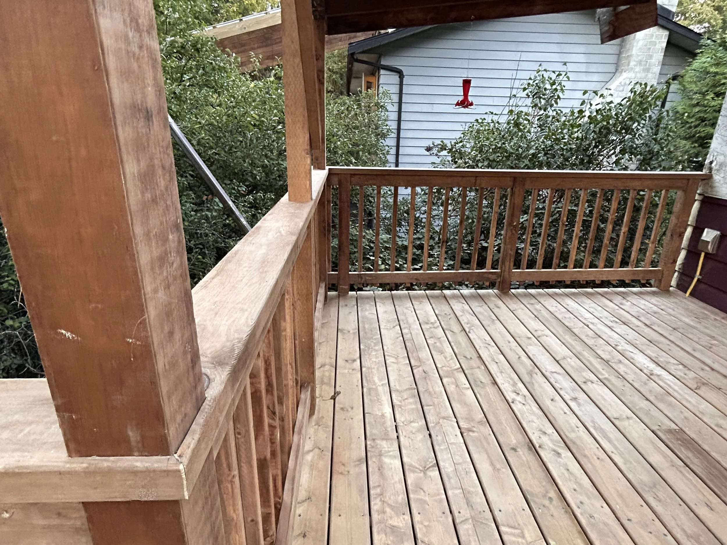 A wooden deck with railing, metal fastener, and surrounding greenery, with a neighboring house and a red hummingbird feeder hanging on the wall in the background.