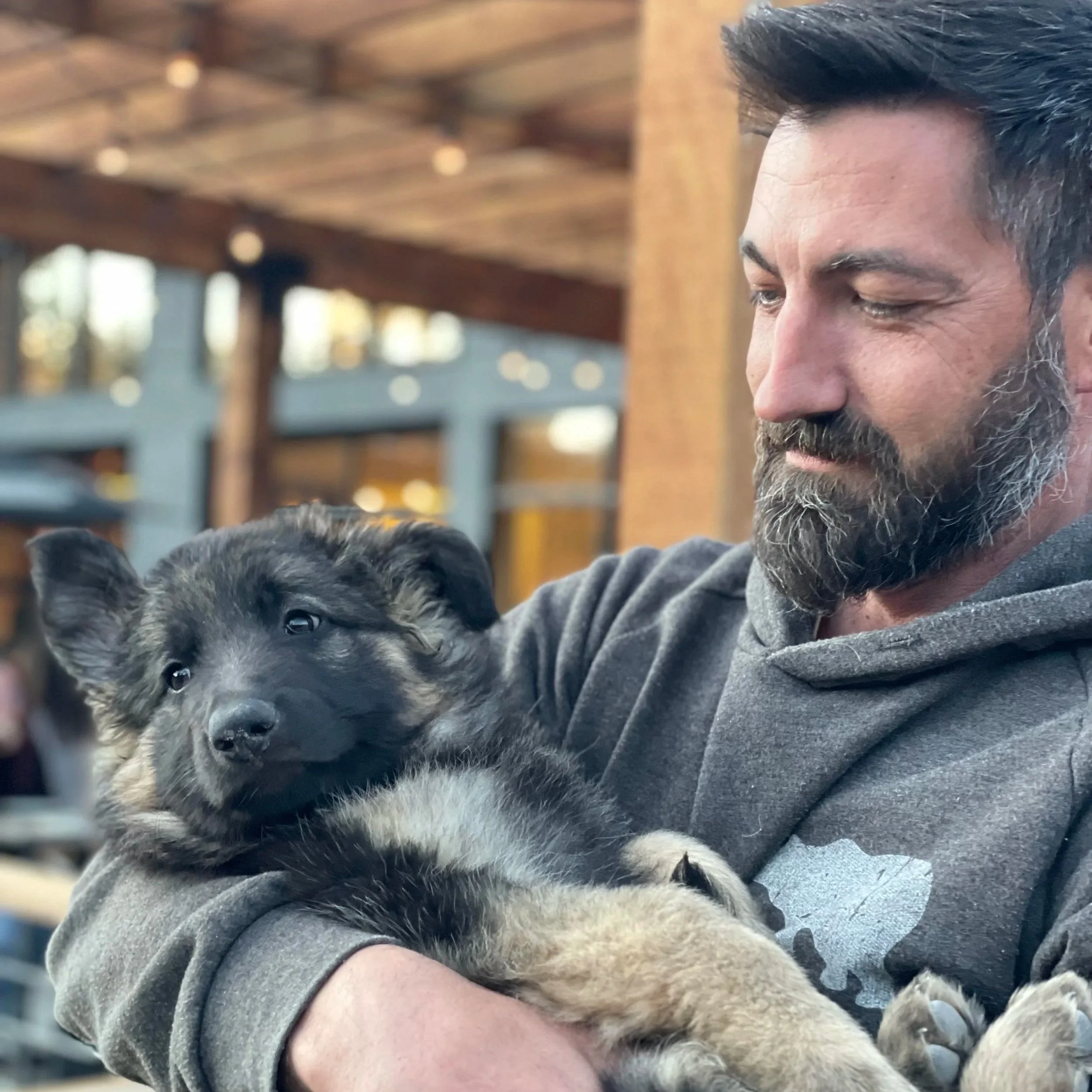 A man holding a black and tan puppy outdoors under a wooden structure.