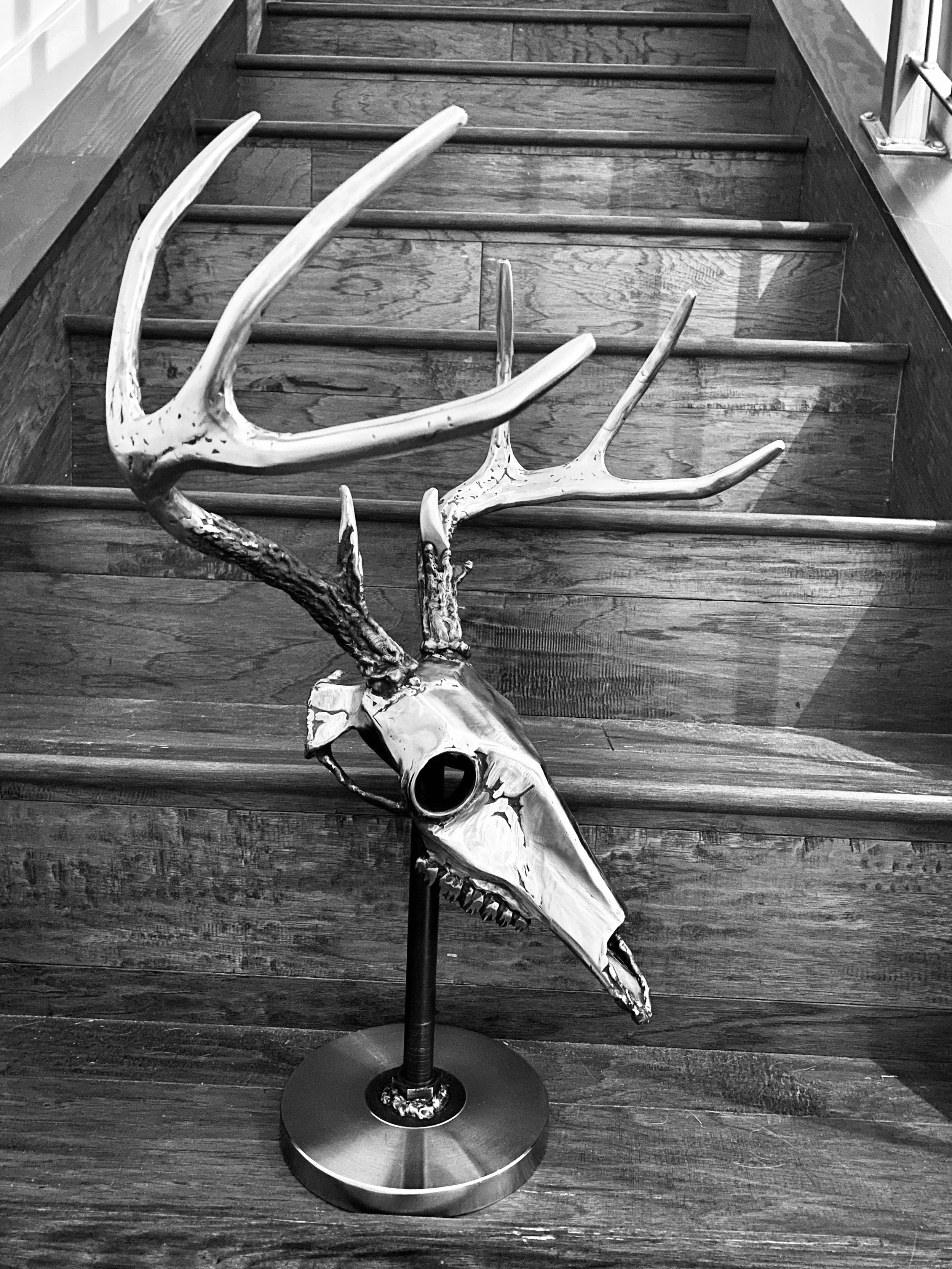 metal sculpture of a deer skull with antlers on a stand, placed on a wooden floor in front of a staircase.