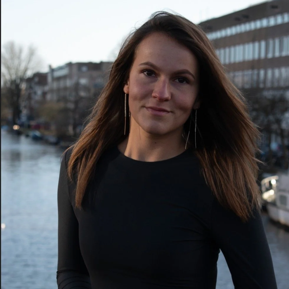 Een vrouw met lange bruin haar, donkere long-sleeve shirt, staand bij een waterweg met boten, in een stedelijke omgeving 's avonds of bij schemer.