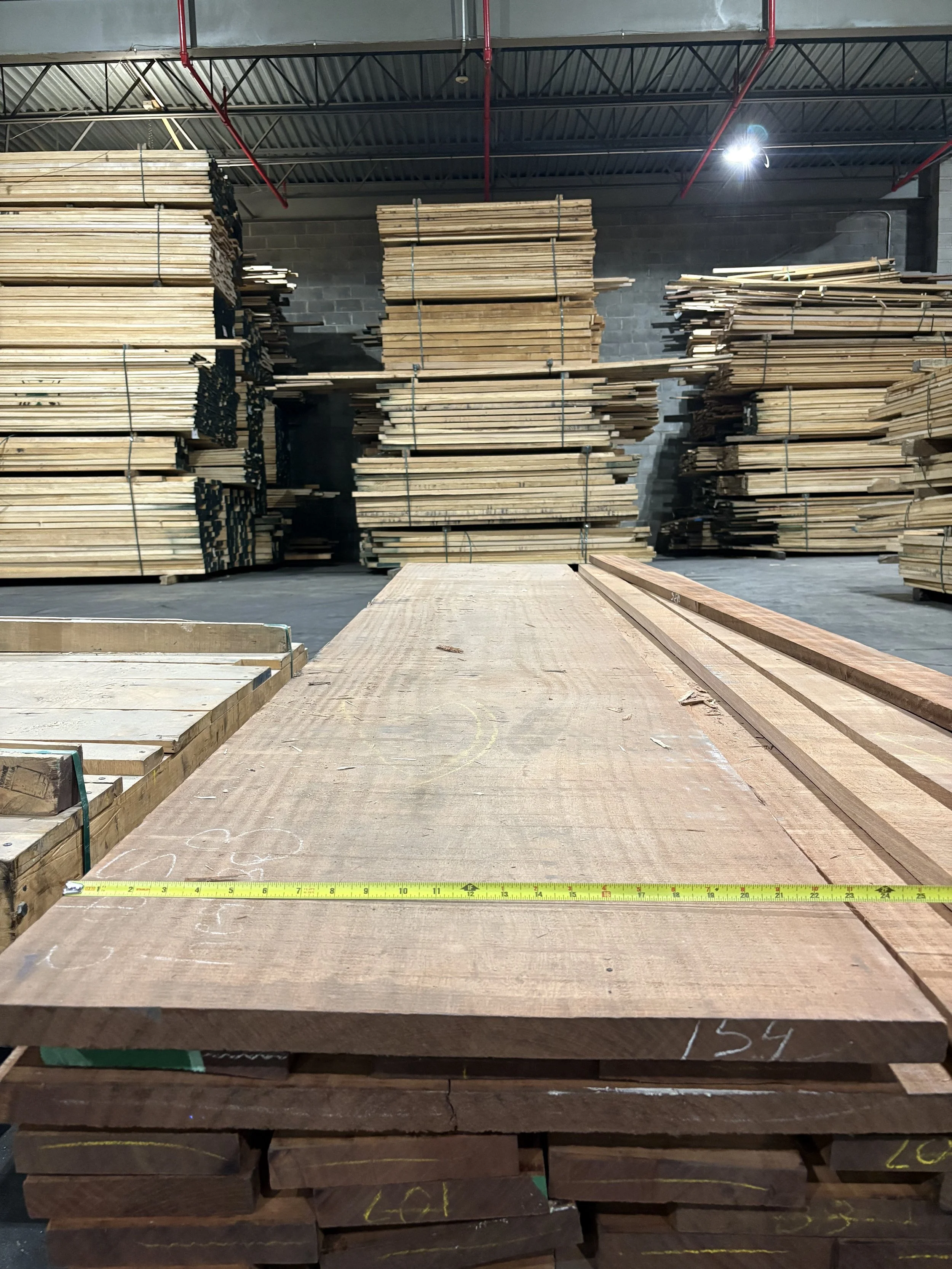 Stacks of lumber and woodworking planks stored in a warehouse with a concrete and brick wall background, and a yellow measuring tape laid across a wooden surface in the foreground.