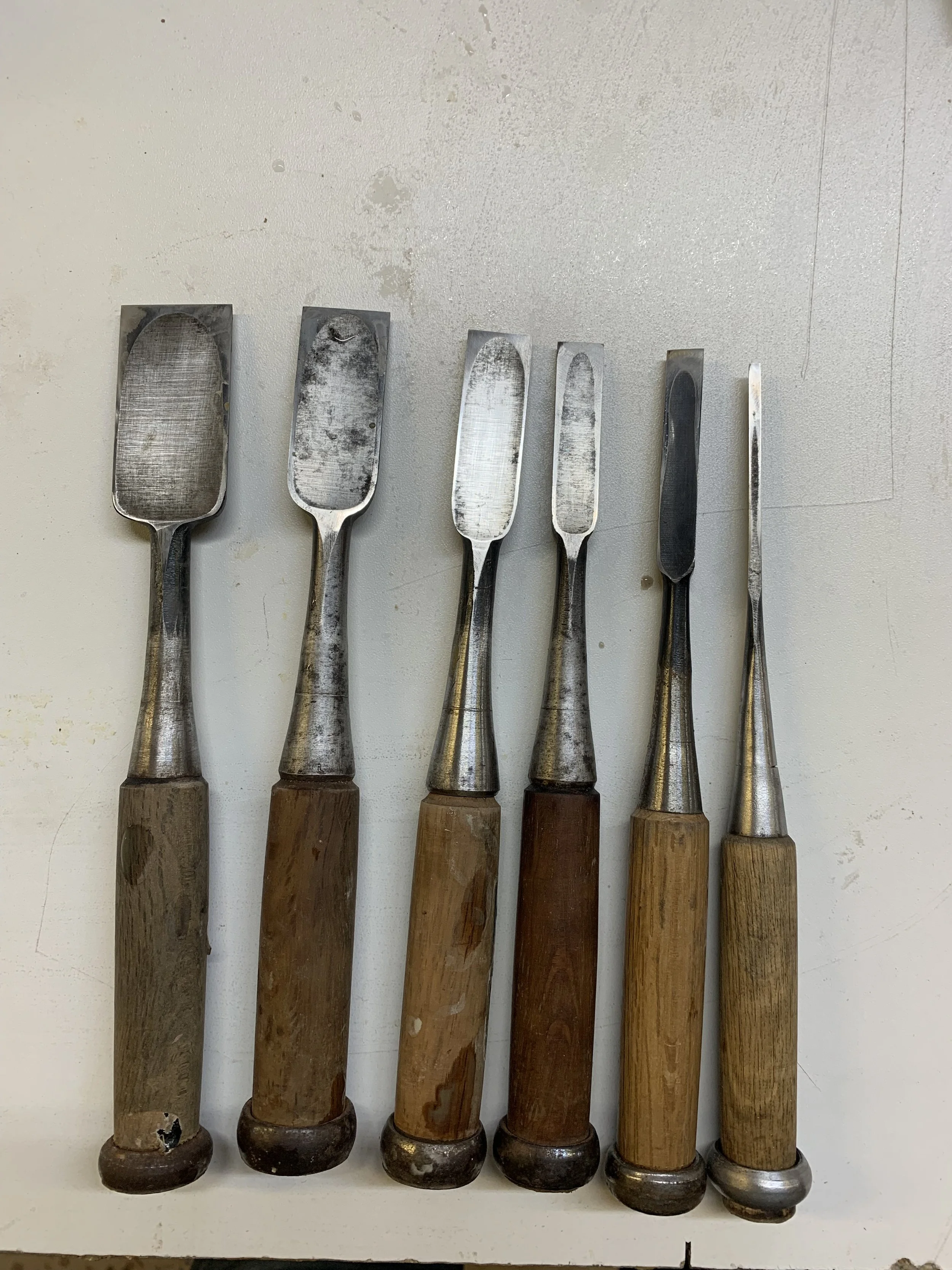 Six vintage metal spatulas with wooden handles arranged in a row on a white surface.