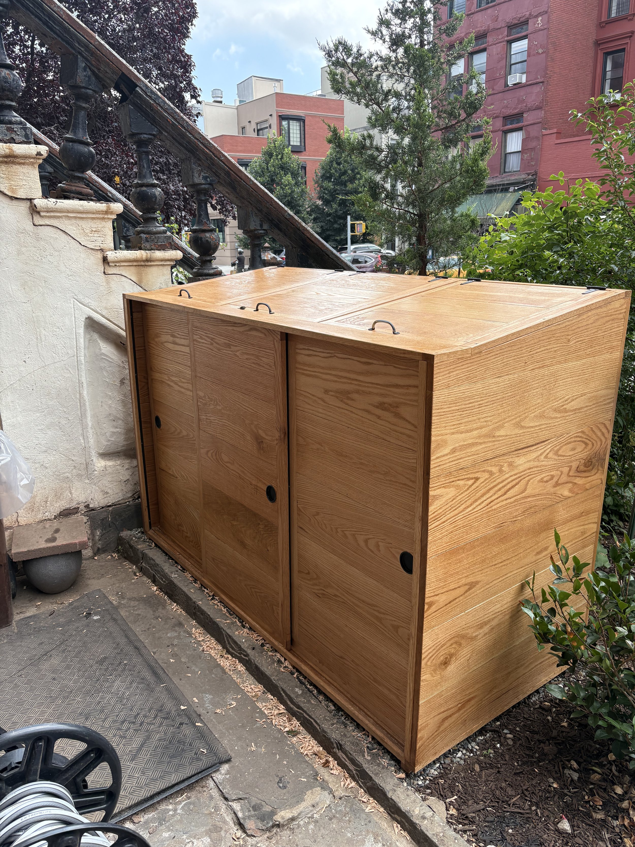 A wooden cabinet with a sliding door is placed outdoors near a building. There is a black metal railing, greenery, and a city street with buildings and cars in the background.