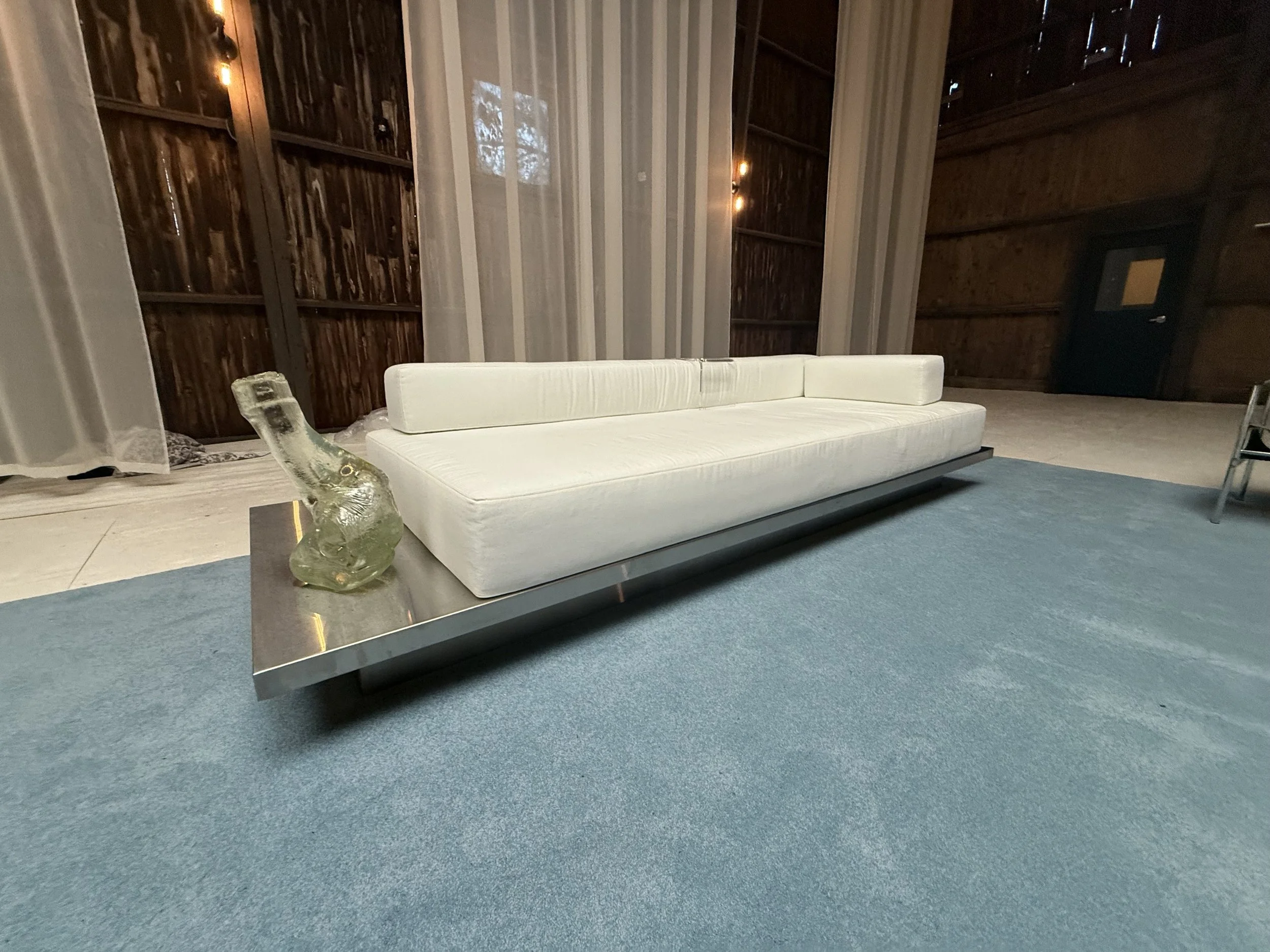 Interior of a wooden building with a cream-colored sofa on a blue rug, a glass horse sculpture on a table, and a dark door at the back. Left side has a white curtain and wood paneling on the wall.
