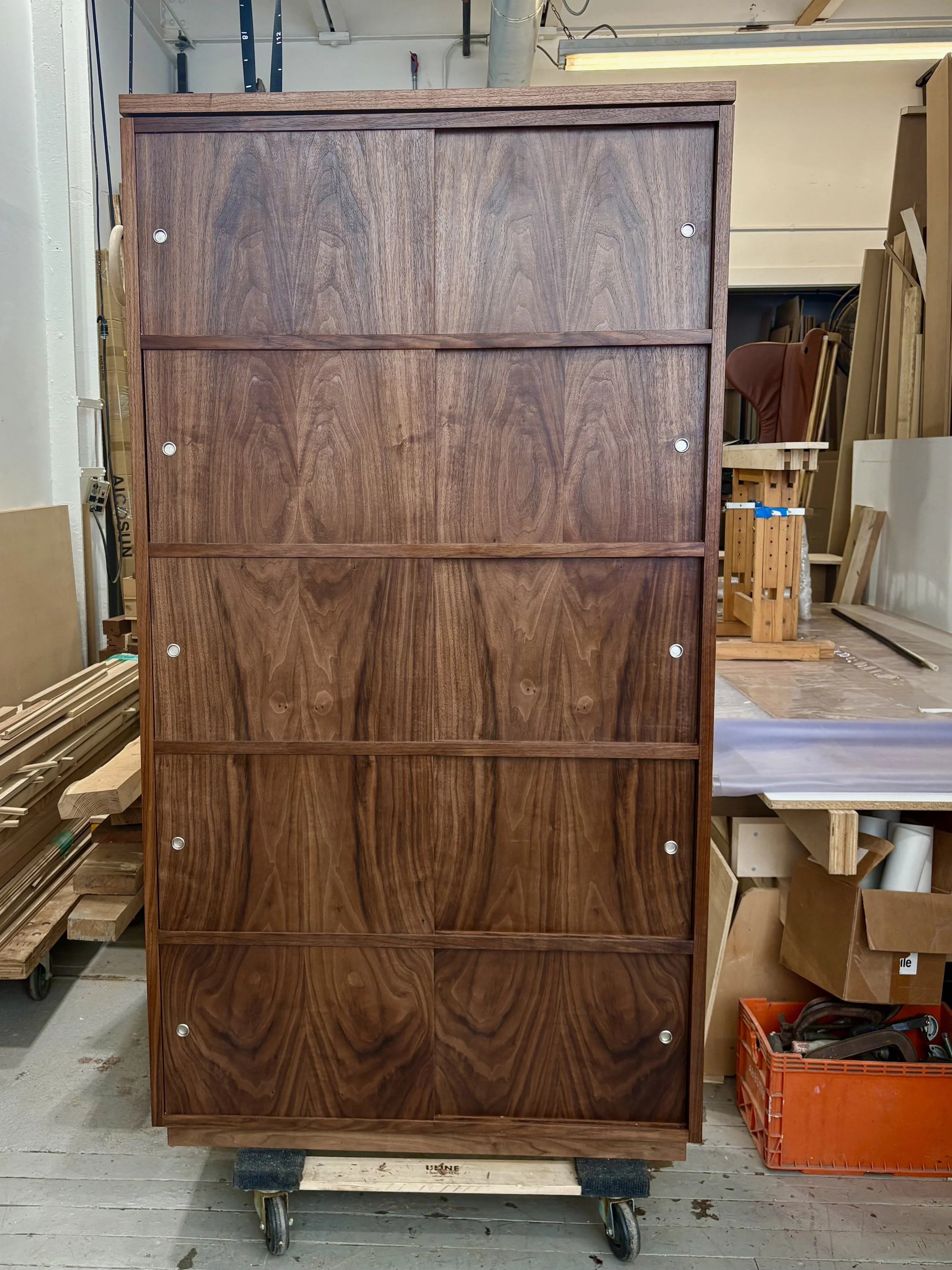 A tall wooden cabinet with a rich wood grain finish, four large drawers, and small circular metal handles. It is placed on a small wheeled platform in a workshop or woodworking space, with various tools and wood pieces visible in the background.