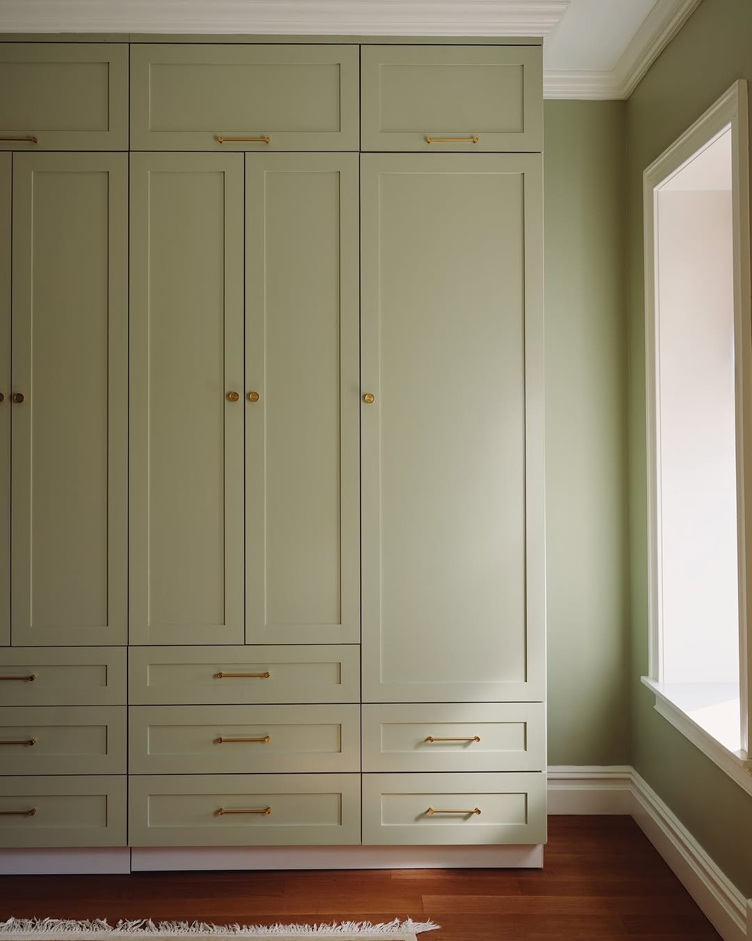 A large built-in green closet with gold hardware located next to a window in a room with green walls and wooden flooring.