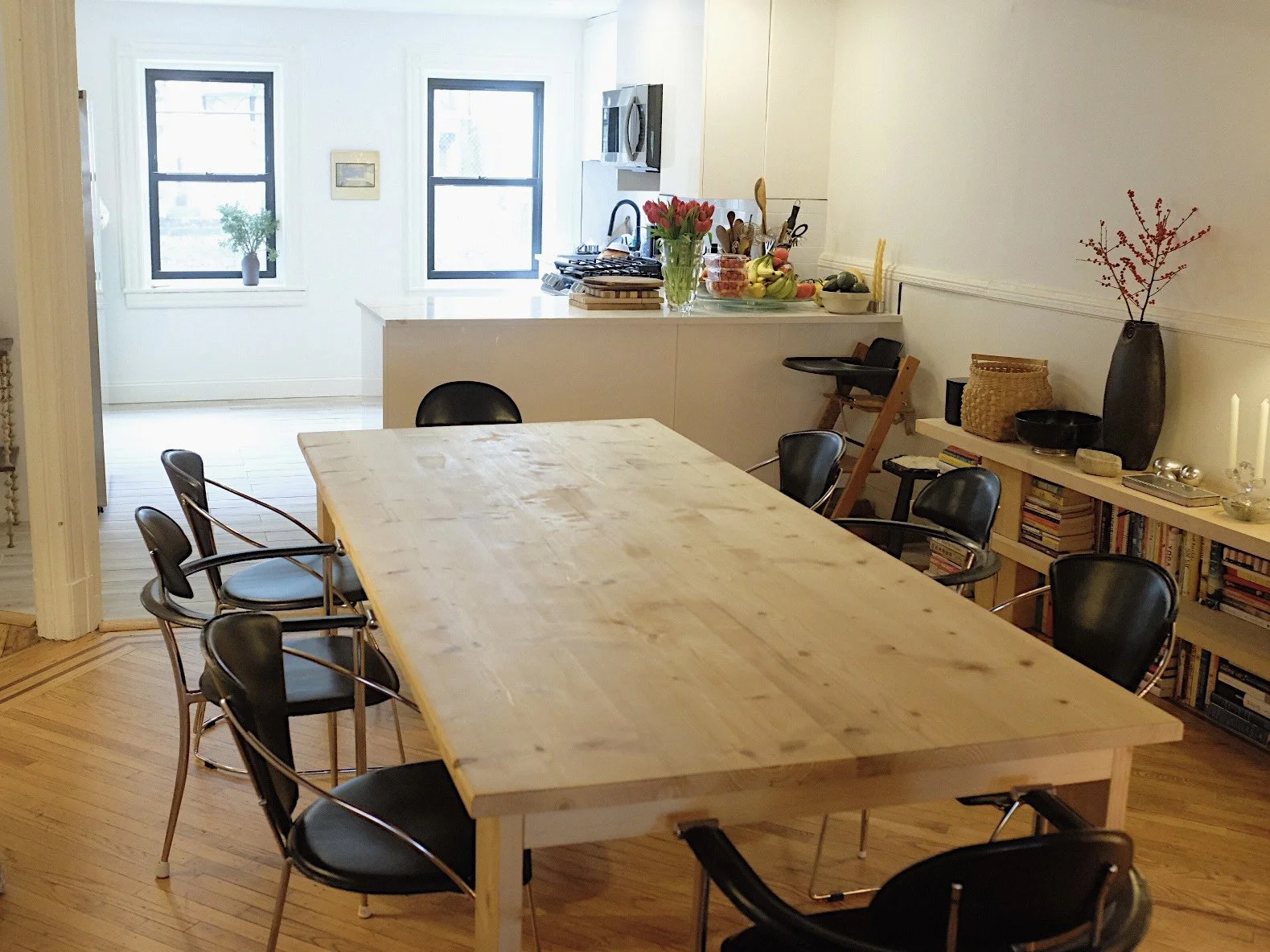 A dining room with a large wooden table surrounded by black chairs and a kitchen with two windows, a countertop with flowers, fruits, and kitchen appliances.