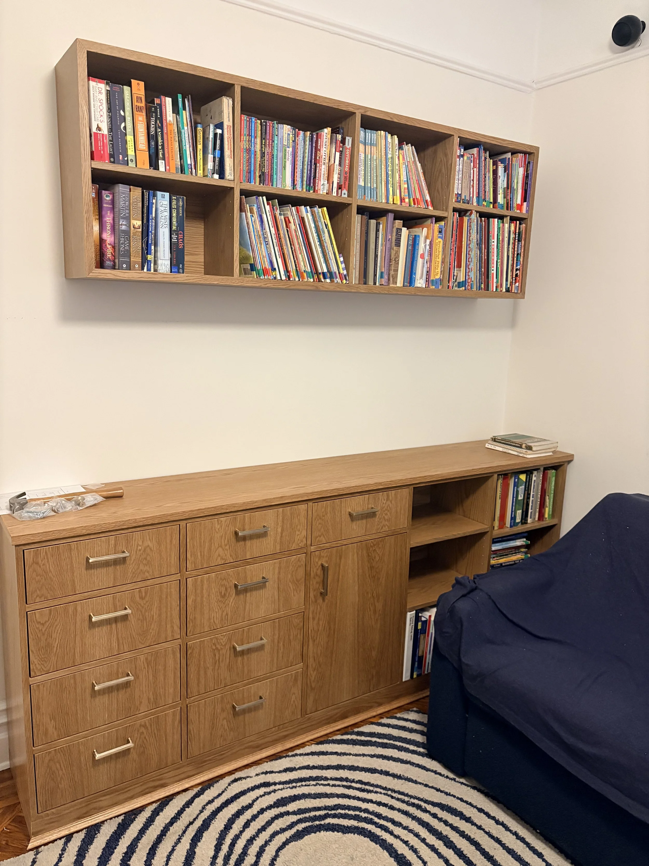 A wooden dresser with multiple drawers and a wooden wall-mounted bookshelf filled with books. A dark blue couch with a blanket on it is partially visible to the right. A rug with black and white concentric circles is on the floor.