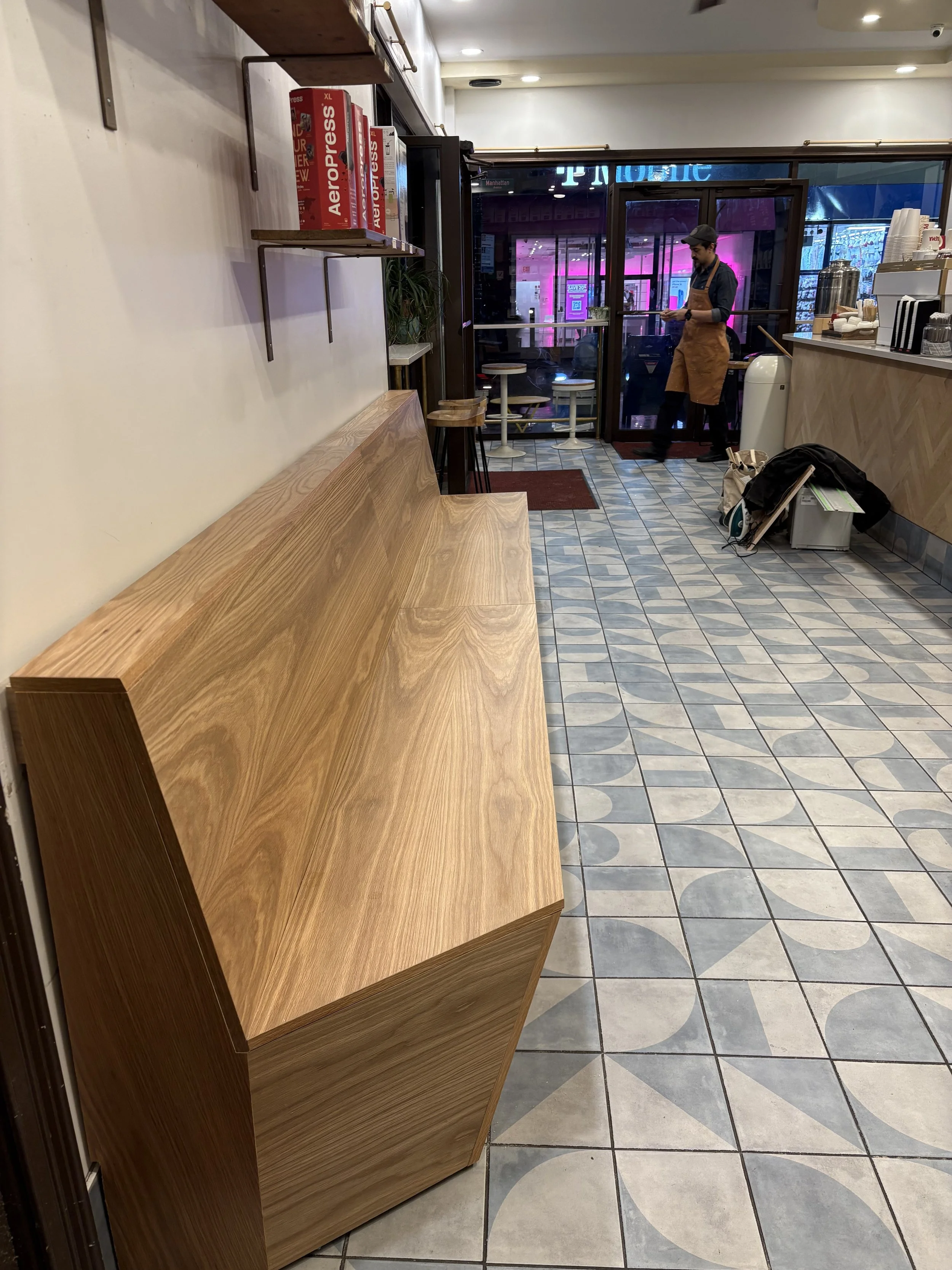 Interior of a cafe with wooden seating, shelves with board games on a wall, and a man in an apron reading a menu near the entrance. Outside neon signs and street view visible through glass door.