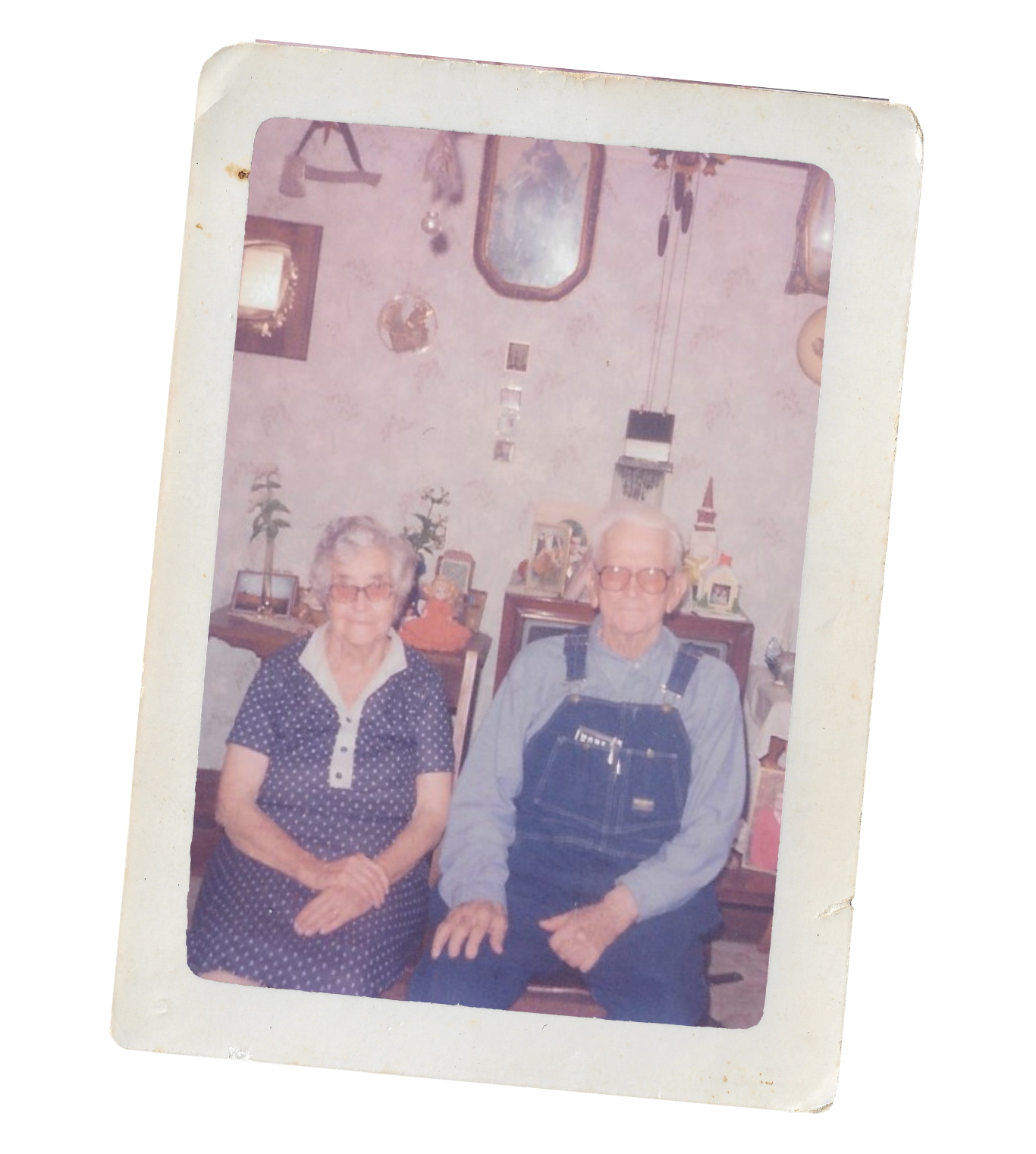 An old photograph of an elderly woman and man sitting next to each other in a living room, surrounded by various decorative items and plants on shelves behind them.