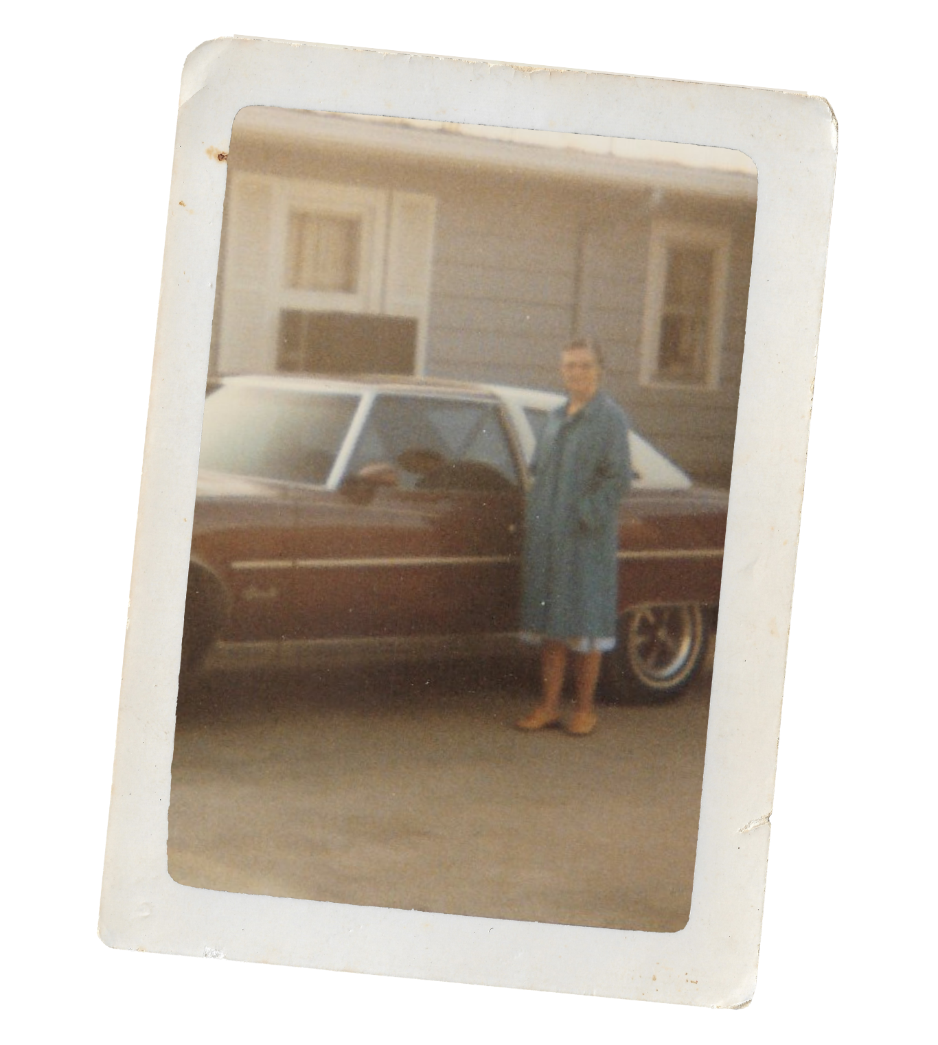 A woman in a long coat and boots standing next to a vintage car in front of a house with wood siding.