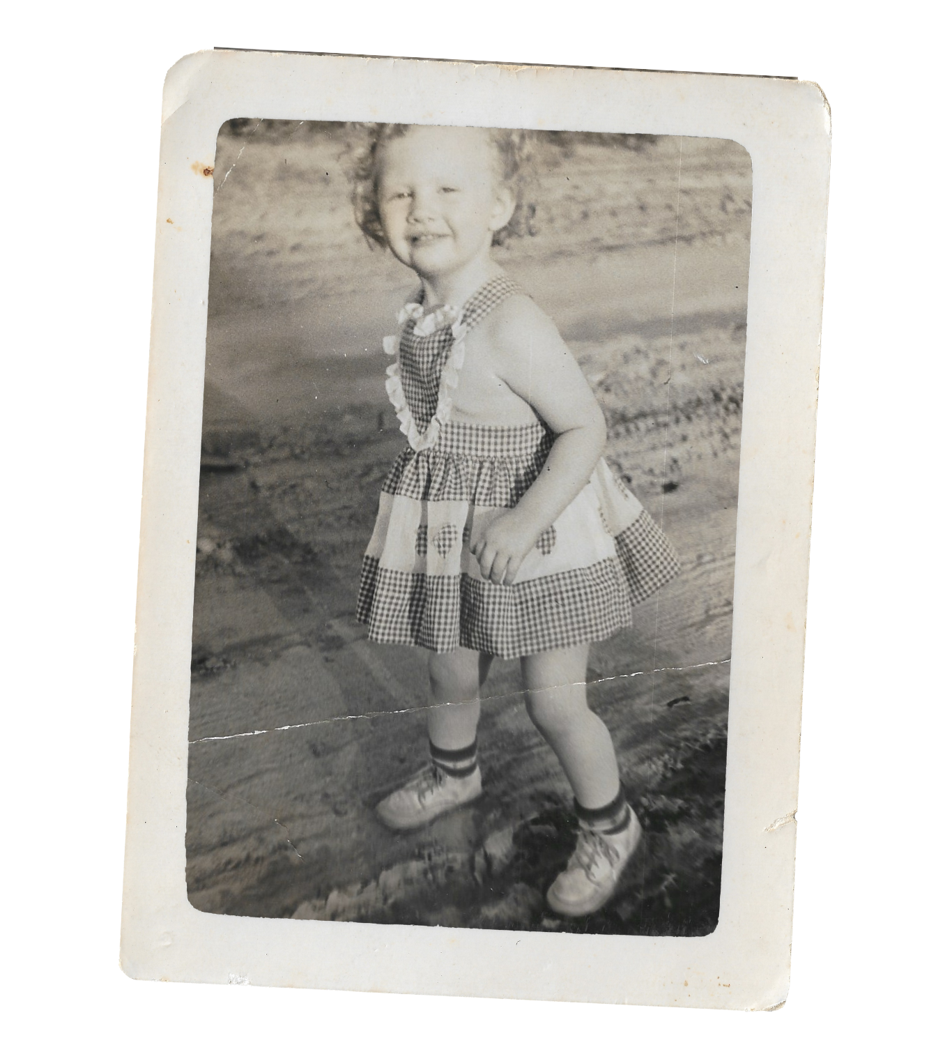 A black and white photograph of a young girl standing outdoors on a dirt path, smiling and posing with her hands on her hips. She is wearing a checkered dress, striped socks, and sneakers, with short curly hair.