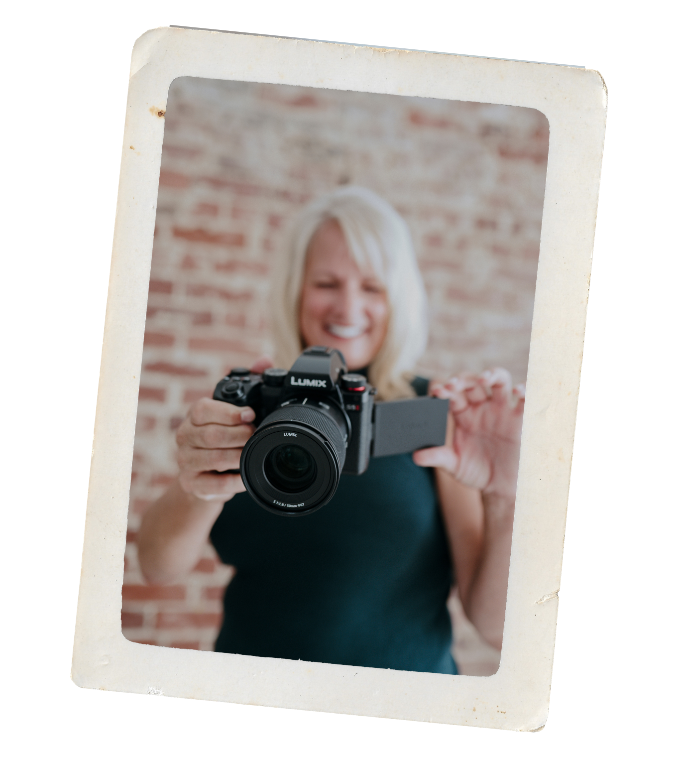 A woman with blonde hair smiling while holding a camera, captured in a Polaroid-style frame against a brick wall background.