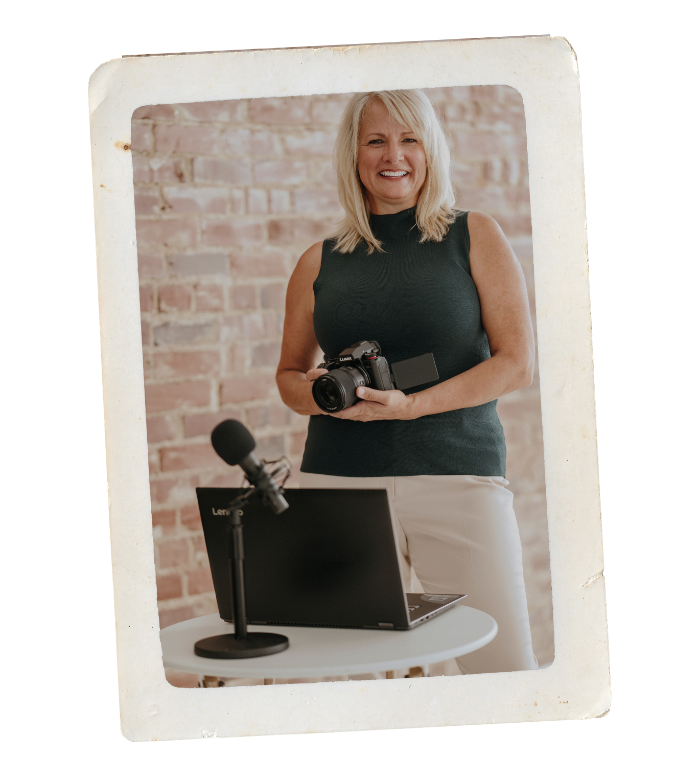 A woman stands in front of a brick wall, holding a camera. She is smiling and wearing a sleeveless dark green top with white pants. On a table in front of her, there is an open laptop, a microphone, and some electronic equipment.