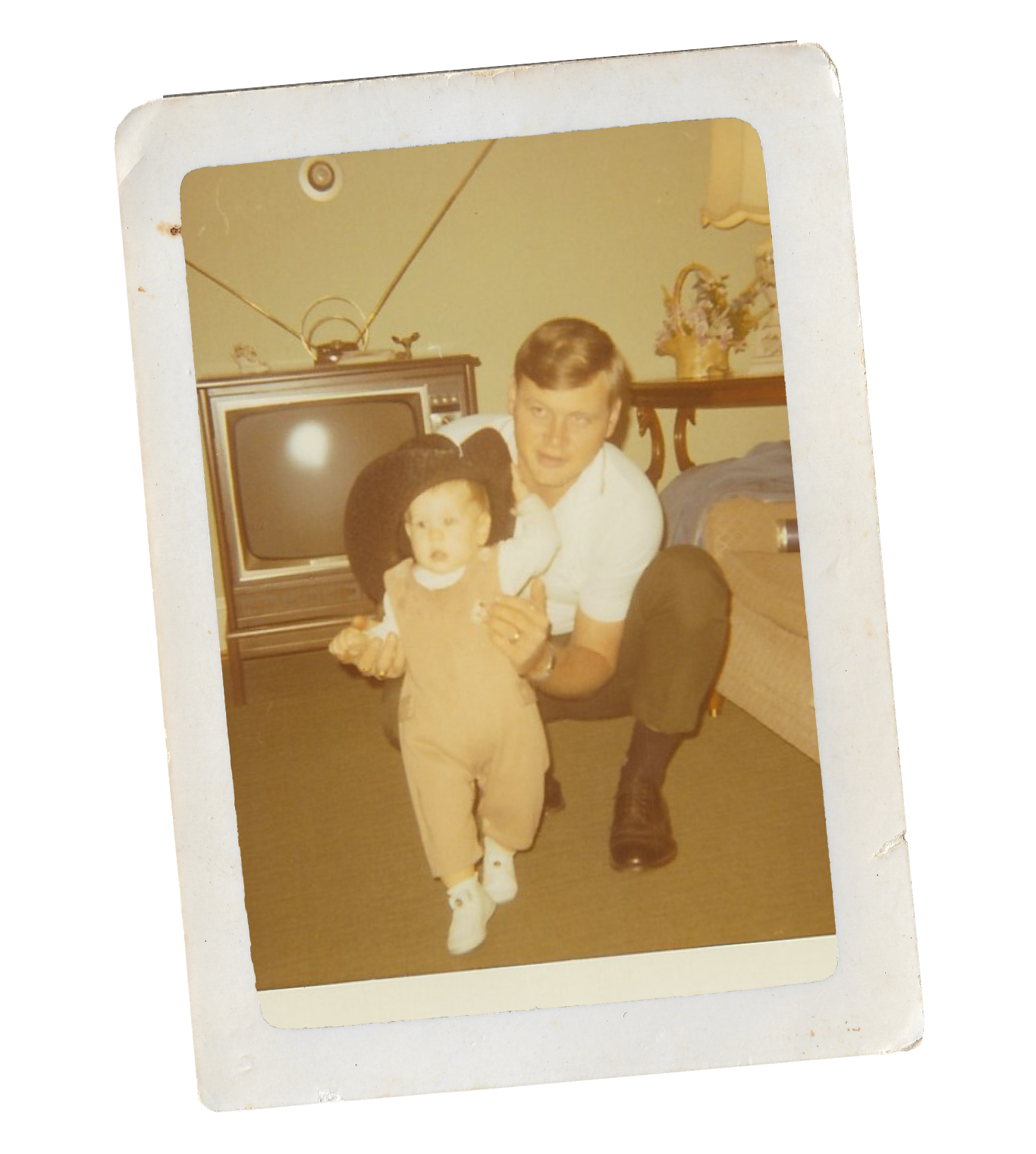 A young man with short hair and a white shirt kneeling beside a small child in a beige jumpsuit and black hat, inside a living room with an old-fashioned TV, a couch, and decorative items on a table.