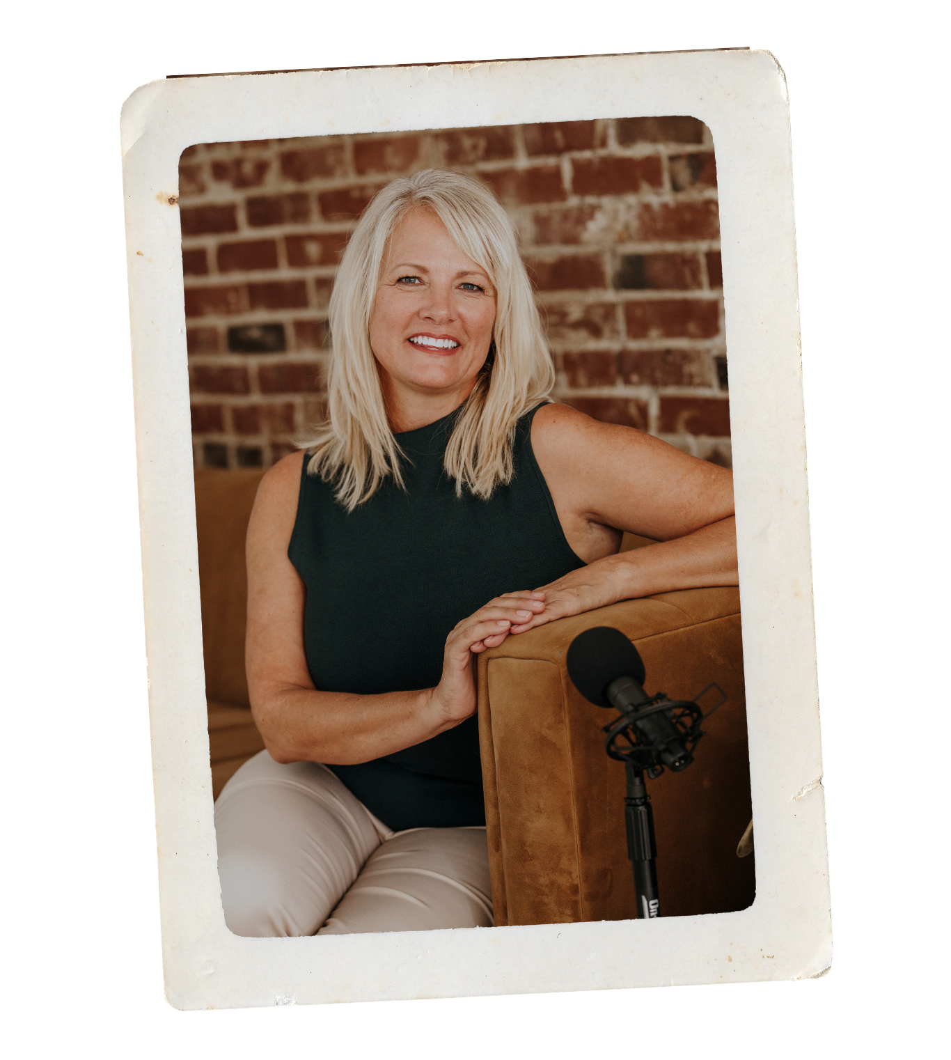 A smiling blonde woman in a black sleeveless top and beige pants sitting on a brown couch, resting her arm on the armrest near a microphone.
