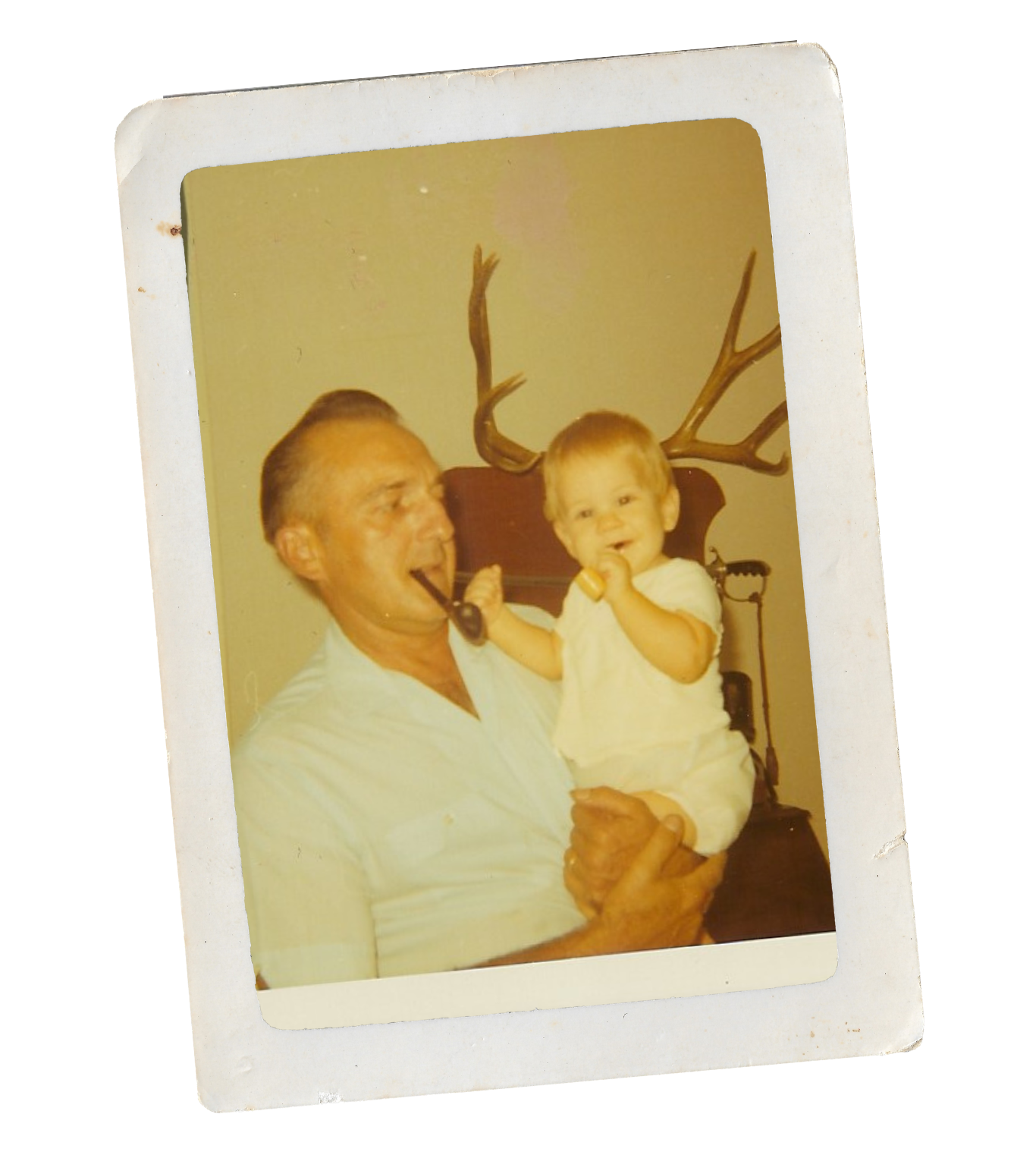 A man holding a young child, both smiling, the man with a pipe in his mouth. A deer antler decor is visible on the wall behind them.