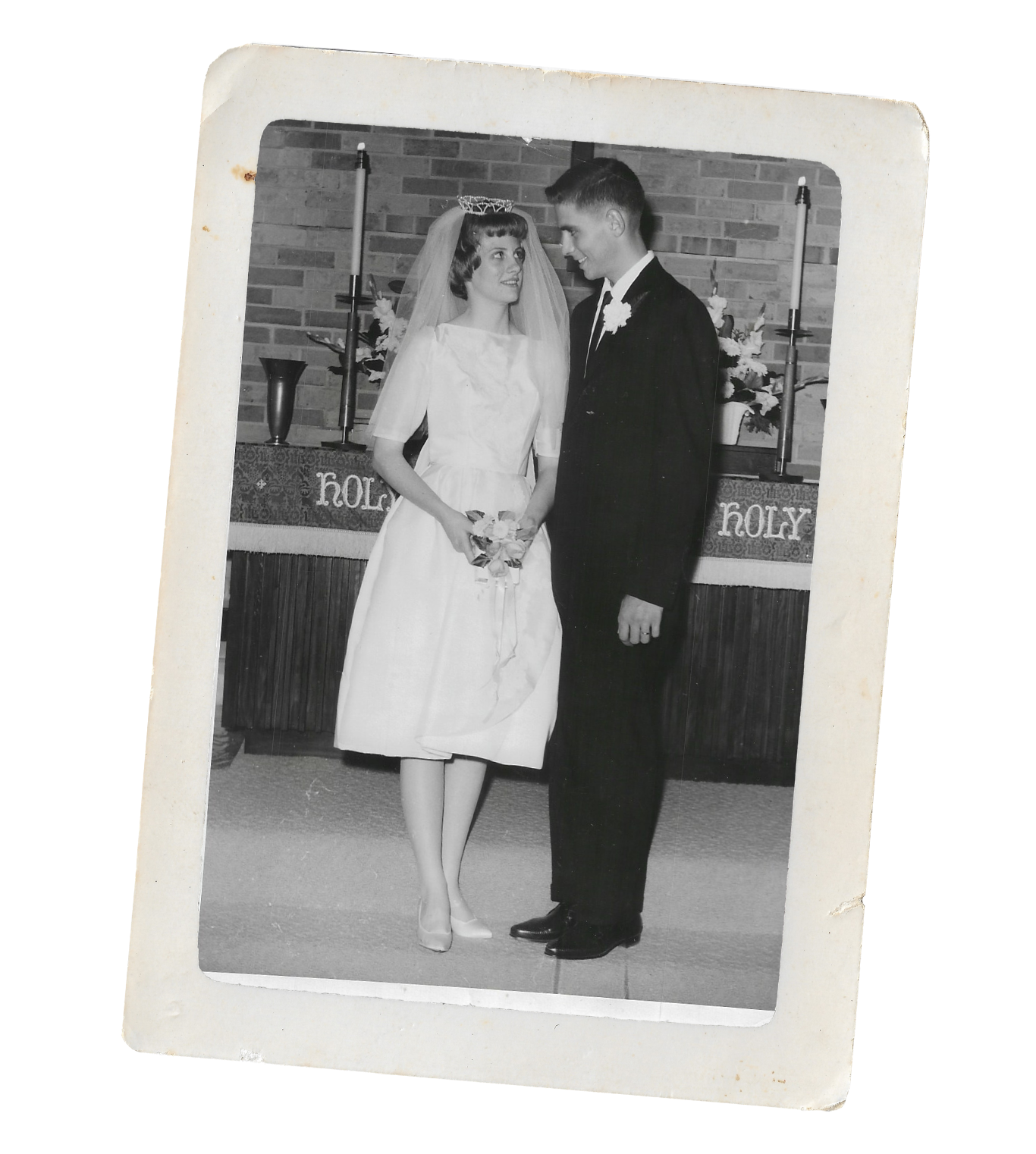 Black and white wedding photograph of a bride and groom standing together indoors, with the bride holding a small bouquet, and a decorated altar with candles and flowers in the background.