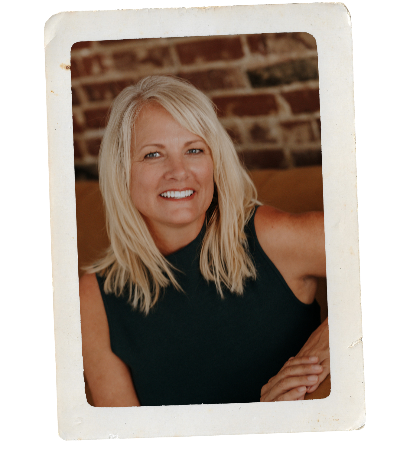 A woman with blonde hair and a black sleeveless top smiling, sitting in front of a brick wall.