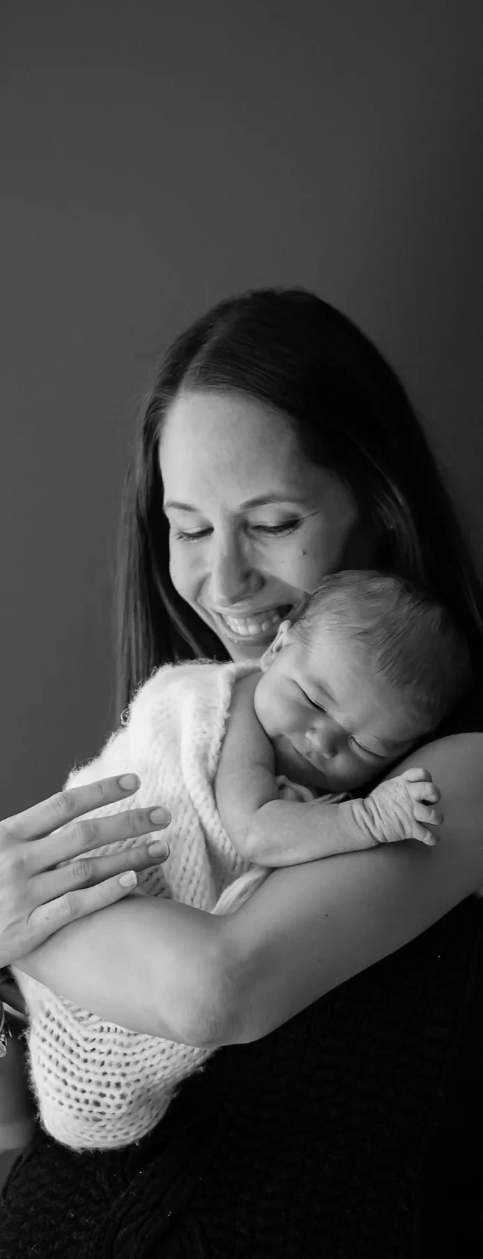 A woman with long hair holding a sleeping baby wrapped in a knitted blanket, smiling softly with her eyes closed, against a plain background.