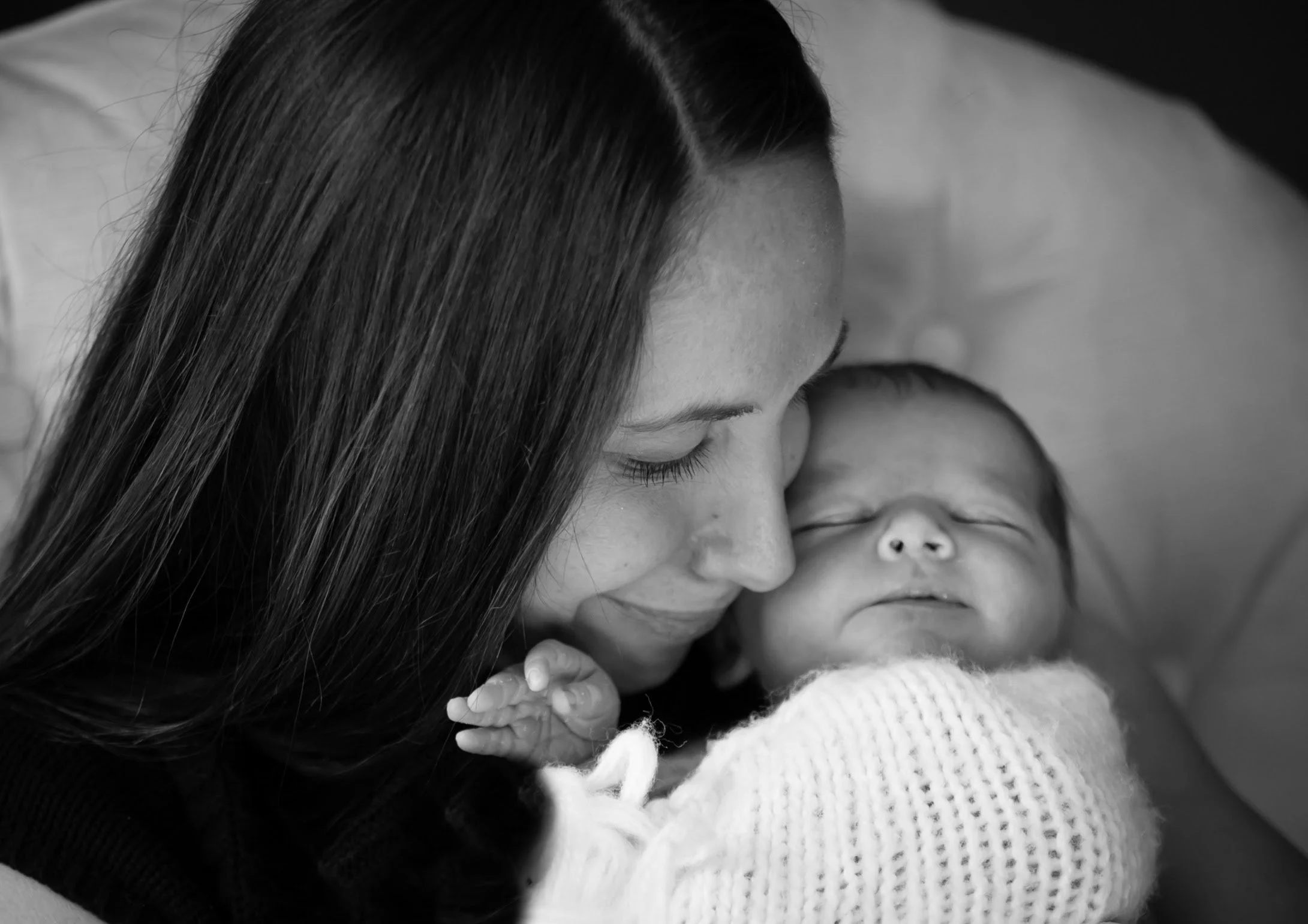 A woman with long dark hair holding a sleeping baby wrapped in a knitted blanket.