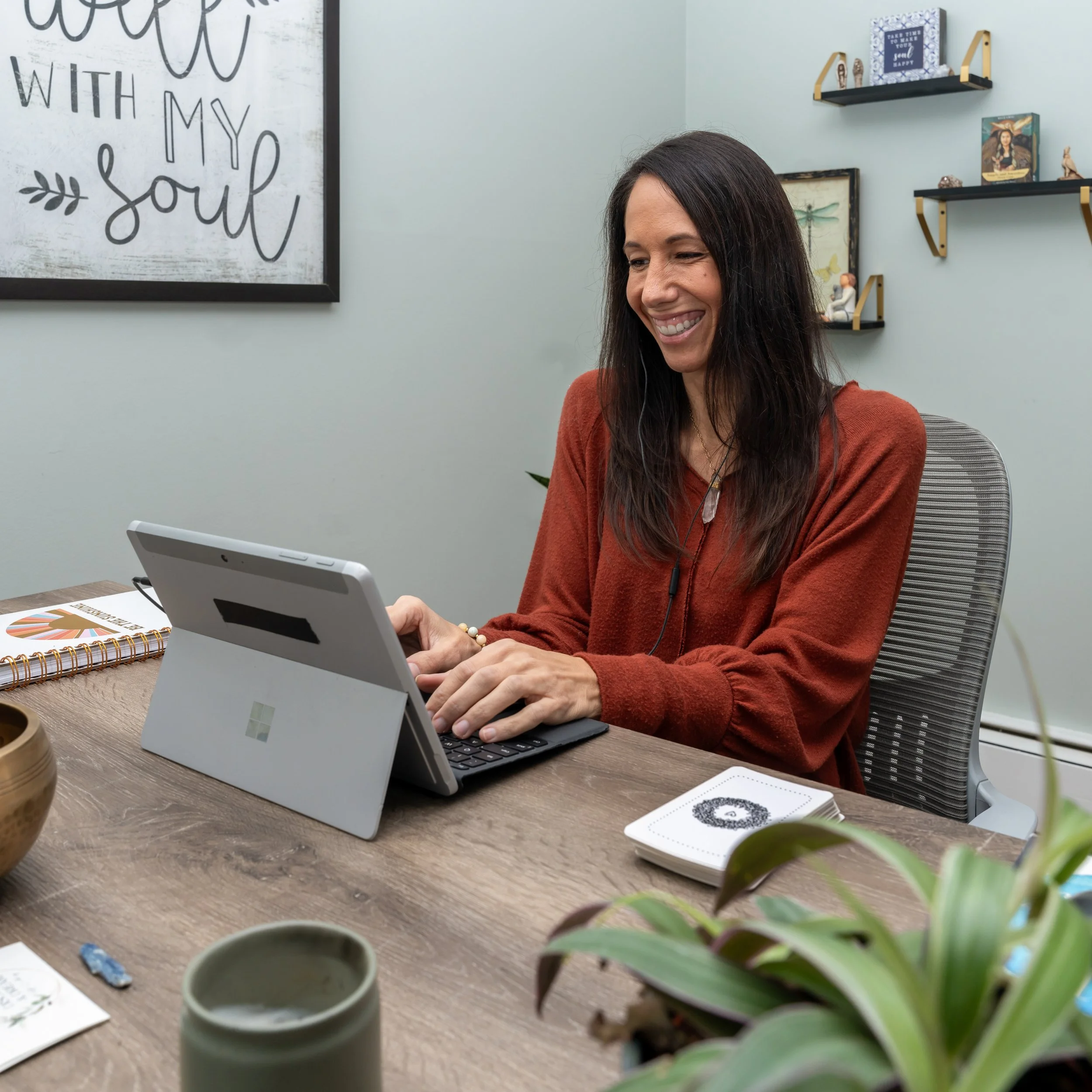 Woman with long brown hair sitting at a wooden desk, smiling while working on a tablet with a keyboard. The room has light gray walls with framed artwork and shelves holding books and decorative items. The desk has various items including a notebook, a small plate with designs, a plant, and a cup.