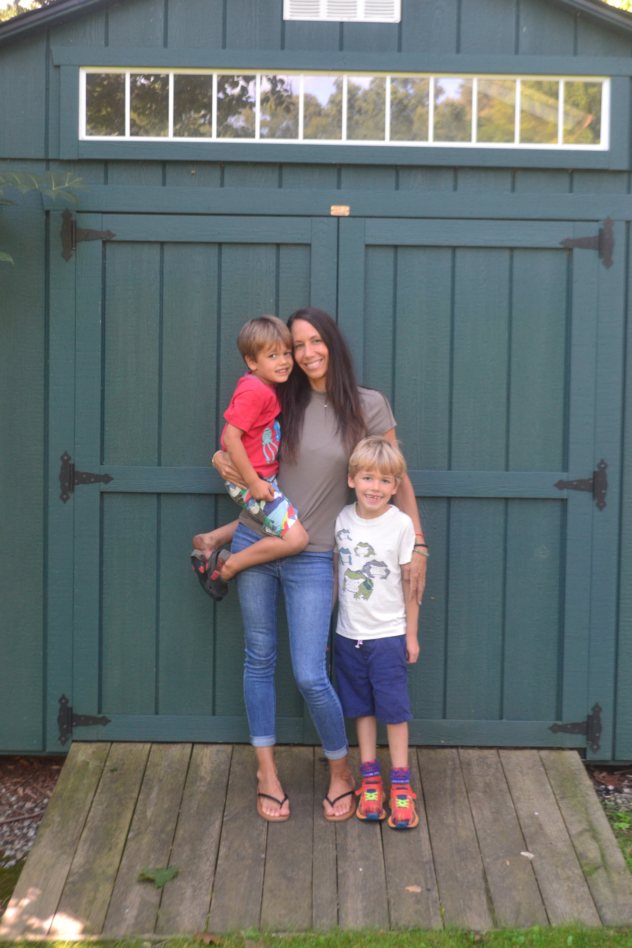 A woman and two young boys standing in front of a green shed, smiling, with one boy being carried.