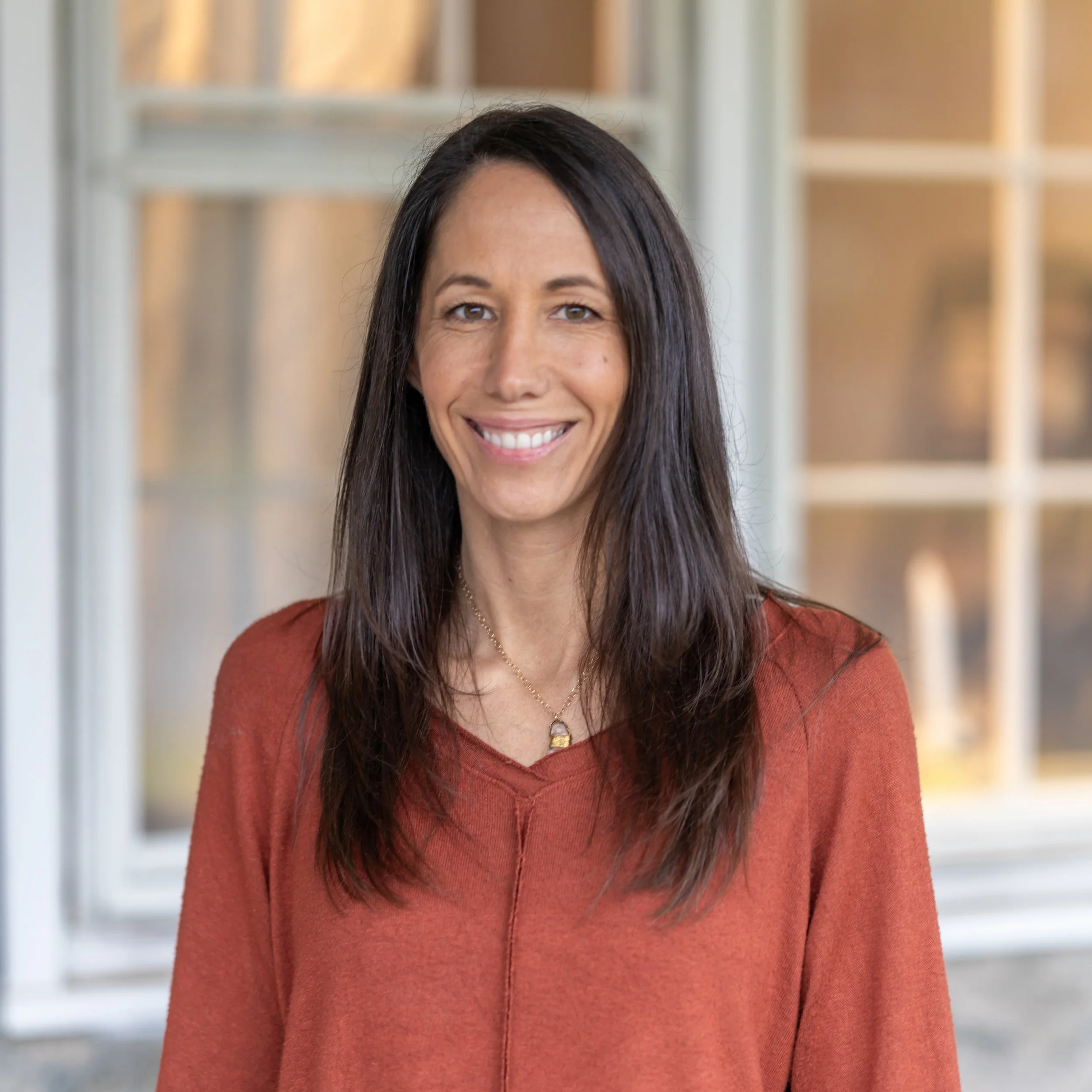 A woman with long dark hair smiling, wearing an orange top and a gold necklace, standing in front of a window.