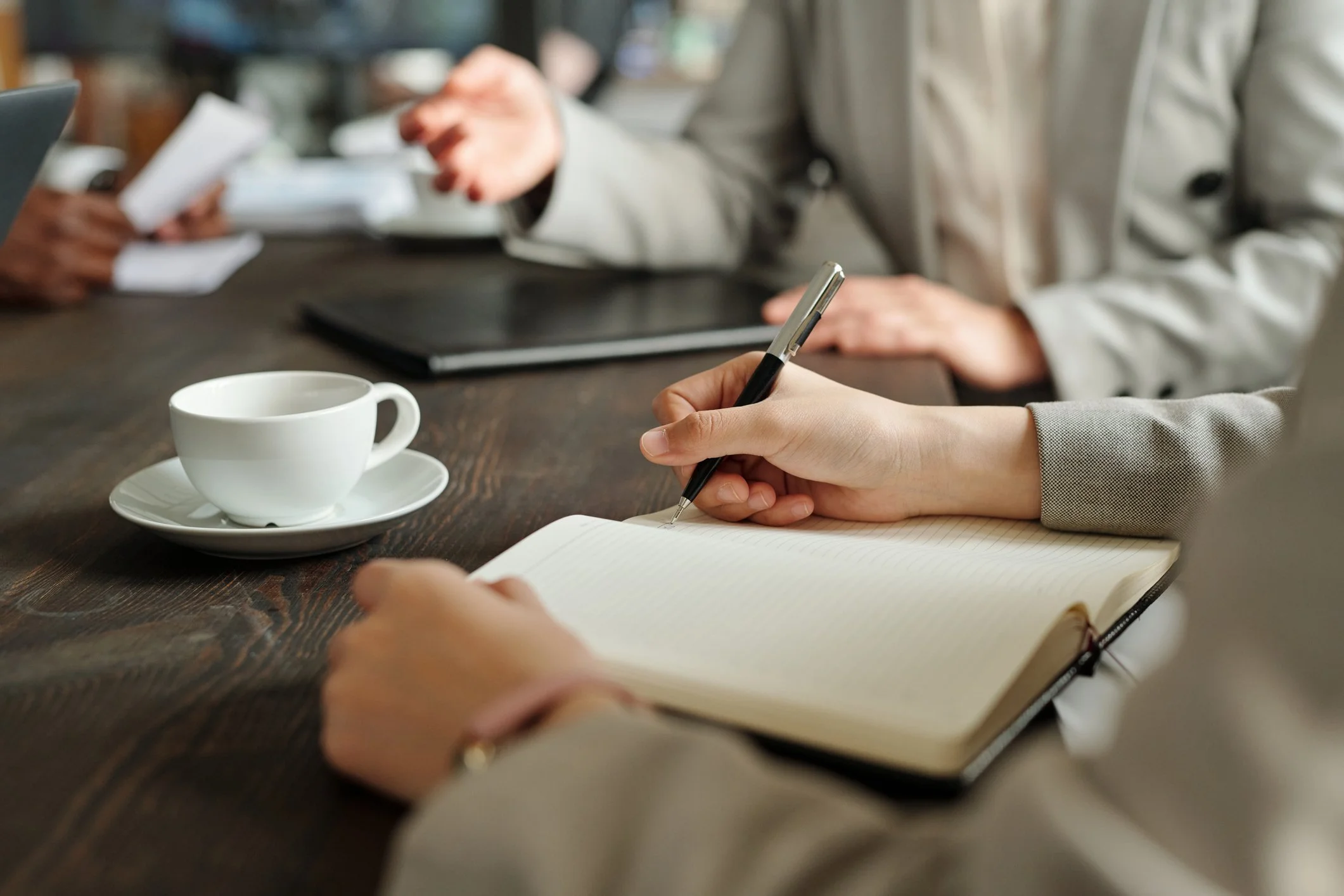Close-up of a person taking notes in a notebook with a pen on a wooden table, near a white coffee cup on a saucer, with other people in the background engaged in conversation or working.