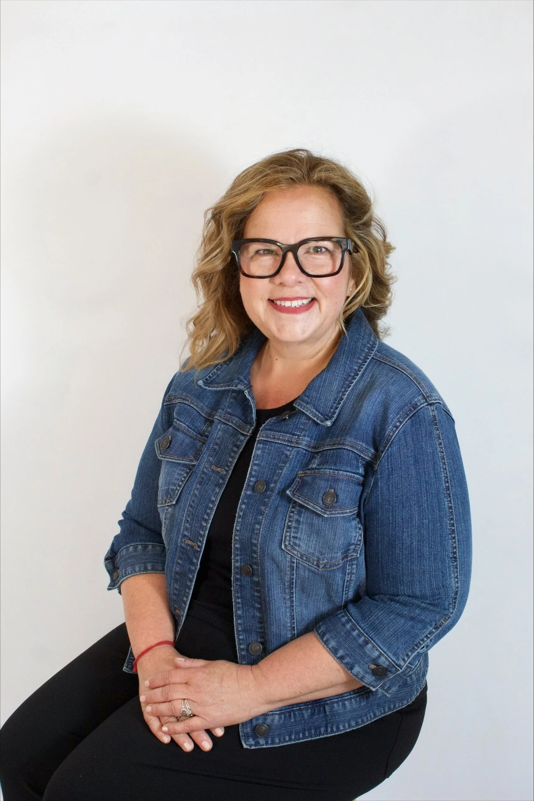 Lisa English, Leadership & Executive Coach and Strategic Consultant,  wearing a denim jacket and black pants, sitting against a white background.