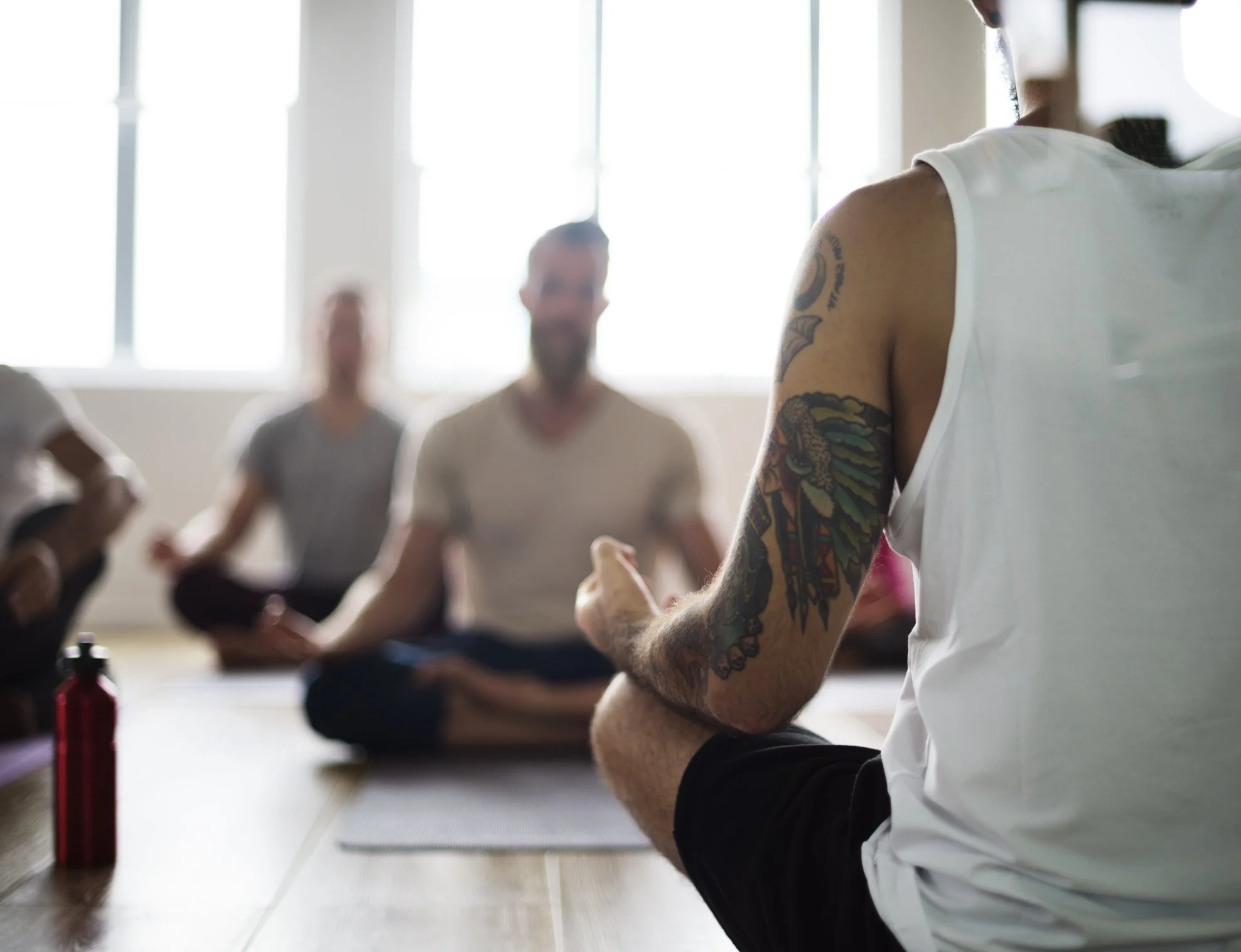 People participating in a yoga class in a bright studio, sitting on mats, with a man in focus showing tattoos on his arm, and others blurred in the background.