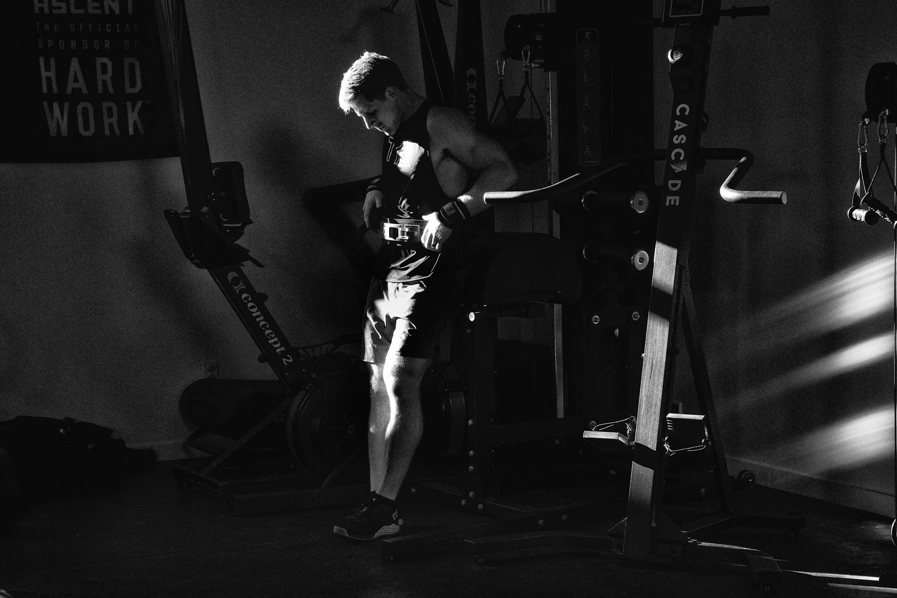 natural light environmental portrait of professional athlete in the shadows to create mood, solemn, black and white. rowing machine, home gym, muscle and shadows