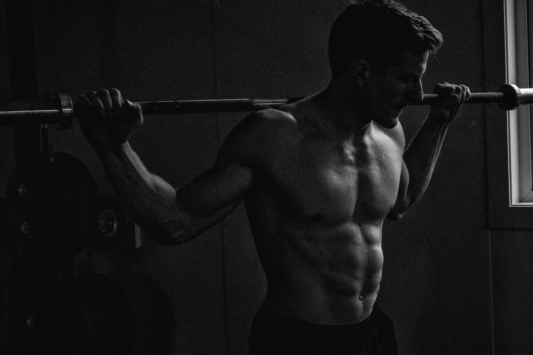 editorial image professional athlete window light  wth barbell on shoulders to create mood and shadow highlighting muscle and 6 pack abs