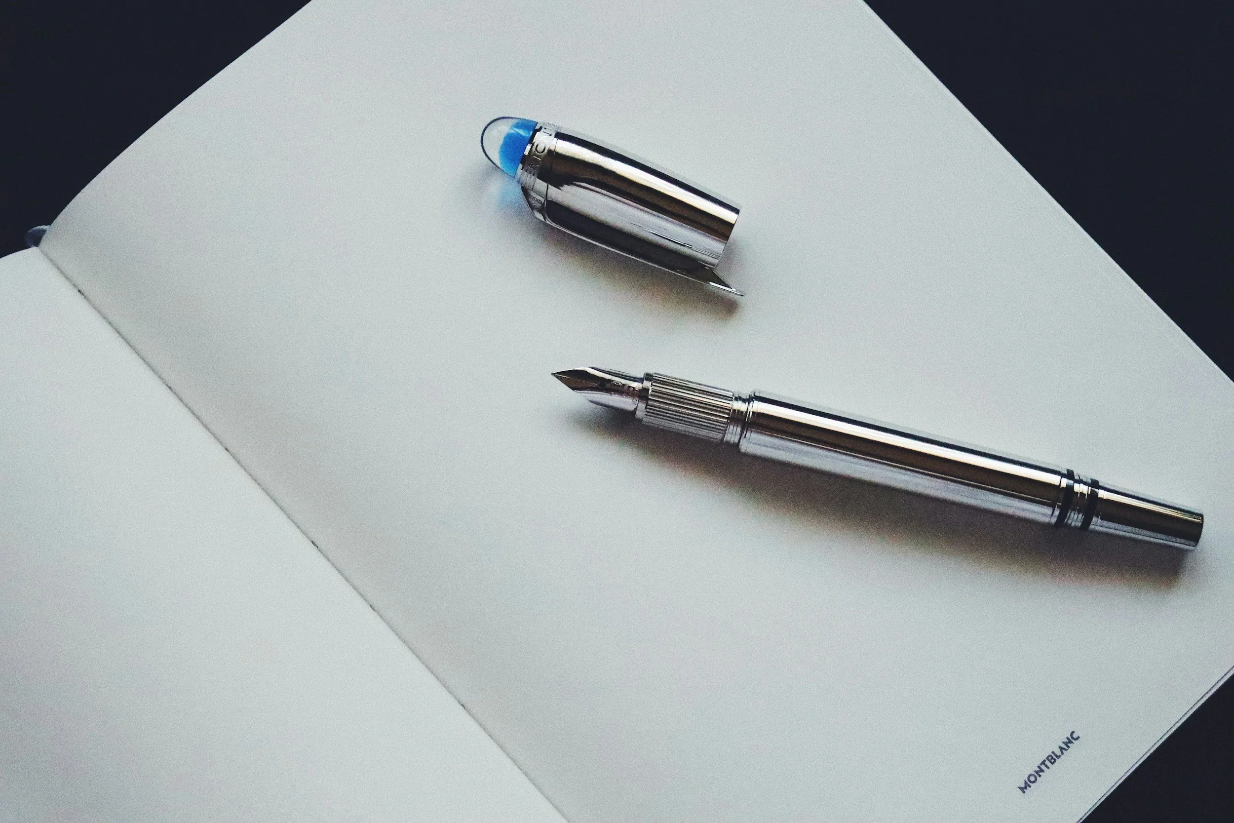 A silver fountain pen with a removable cap lying on an open white notebook with a small Montblanc logo in the bottom corner.