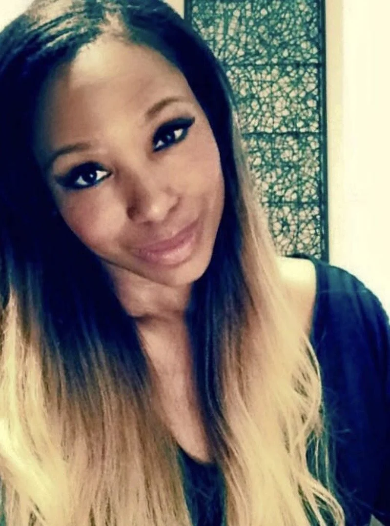 A young woman with long, ombre hair smiling, standing indoors in front of decorative wall art.