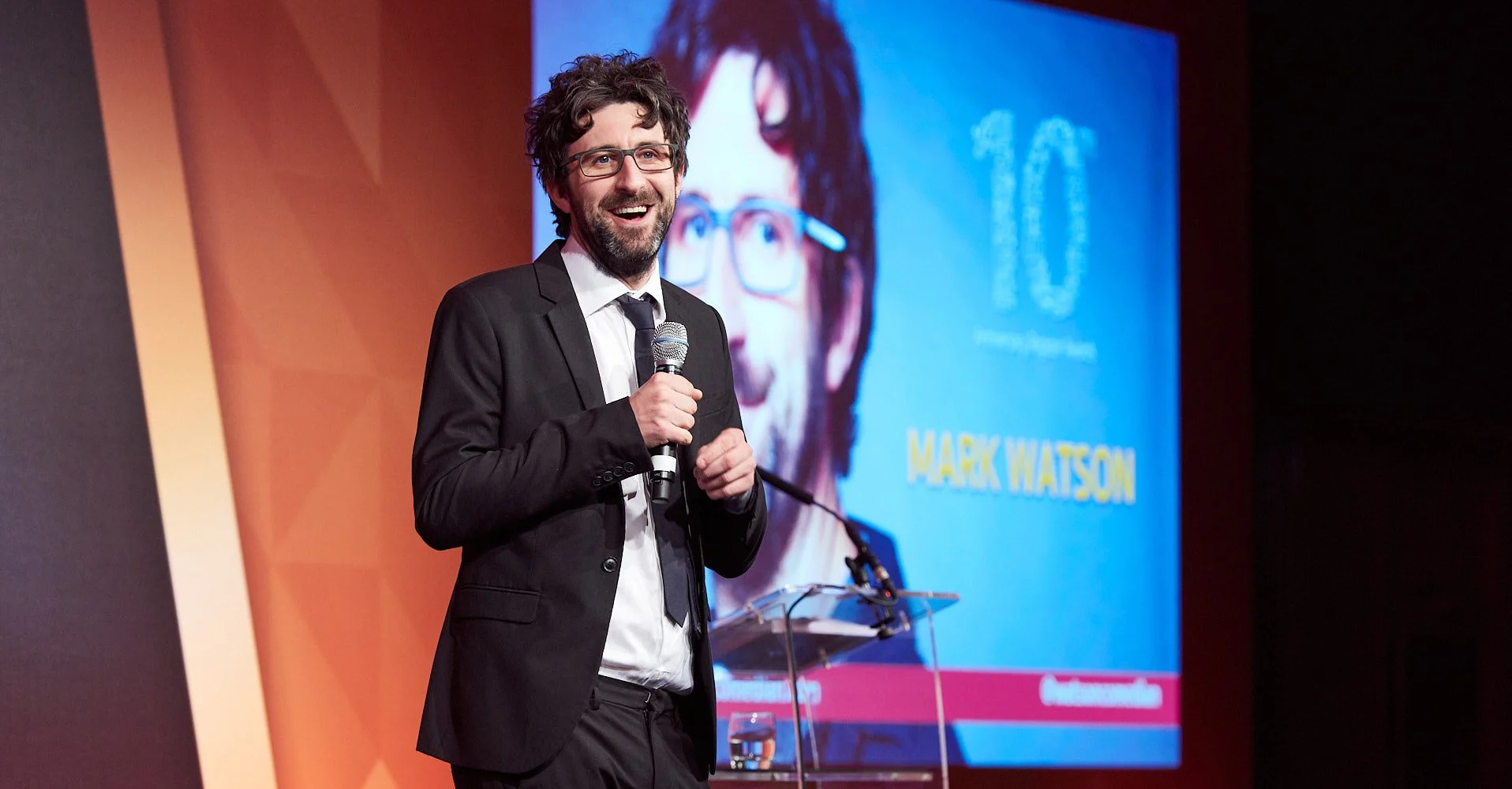 Corporate live event with speaker on illuminated stage comedian Mark Watson