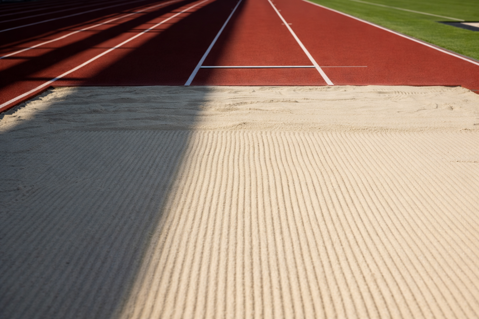 A long outdoor running track with a sandpit in the foreground for long jump or pole vault, and green grass and trees in the background.