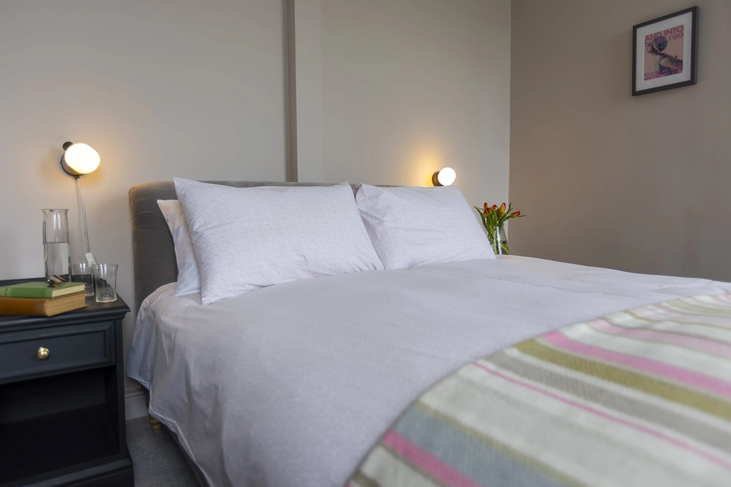 A neatly made bed with white sheets and pillows, a dark nightstand with books, a glass of water, and a decanter, a vase with red and orange flowers, decorated walls with framed art, modern wall-mounted lamps, and a striped blanket at the foot of the bed.