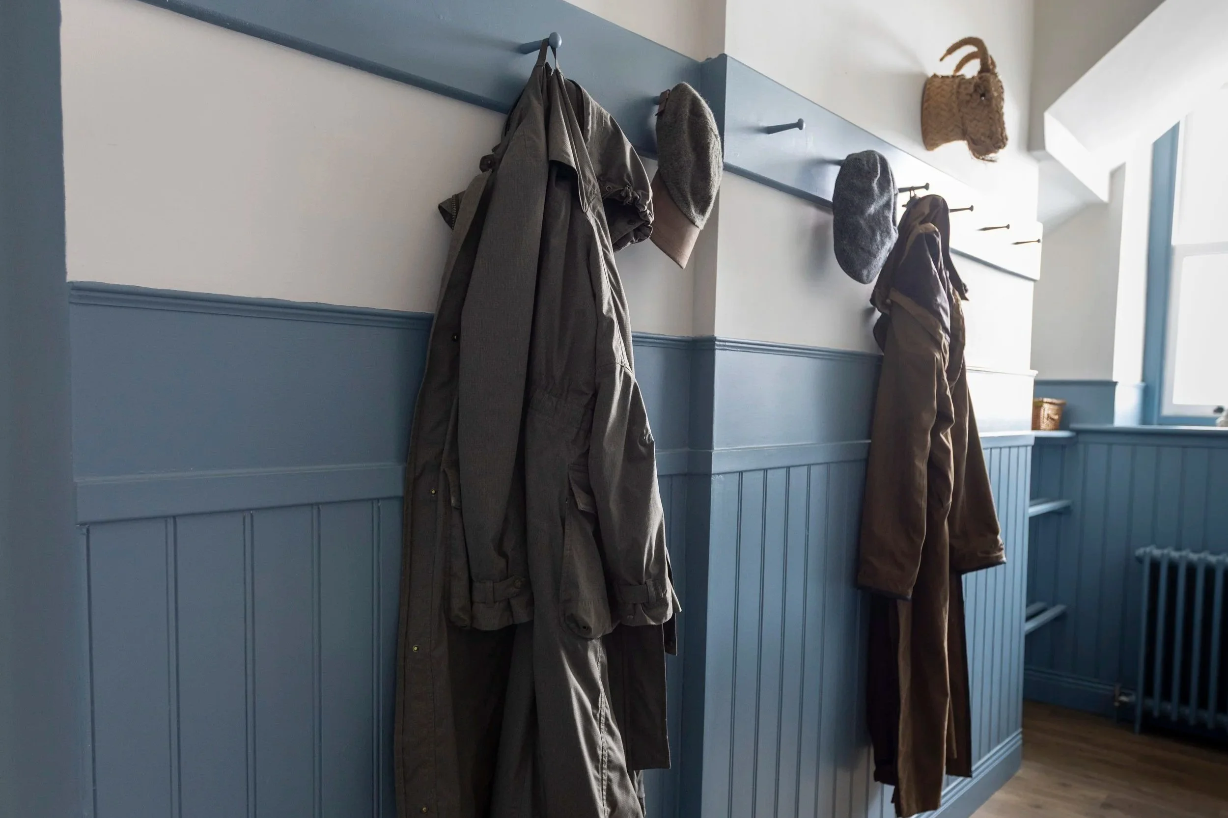 A hallway with blue wainscoting, white walls, and a window. Two coat hooks hold coats and hats, and a pair of black rain boots is on the wooden floor.