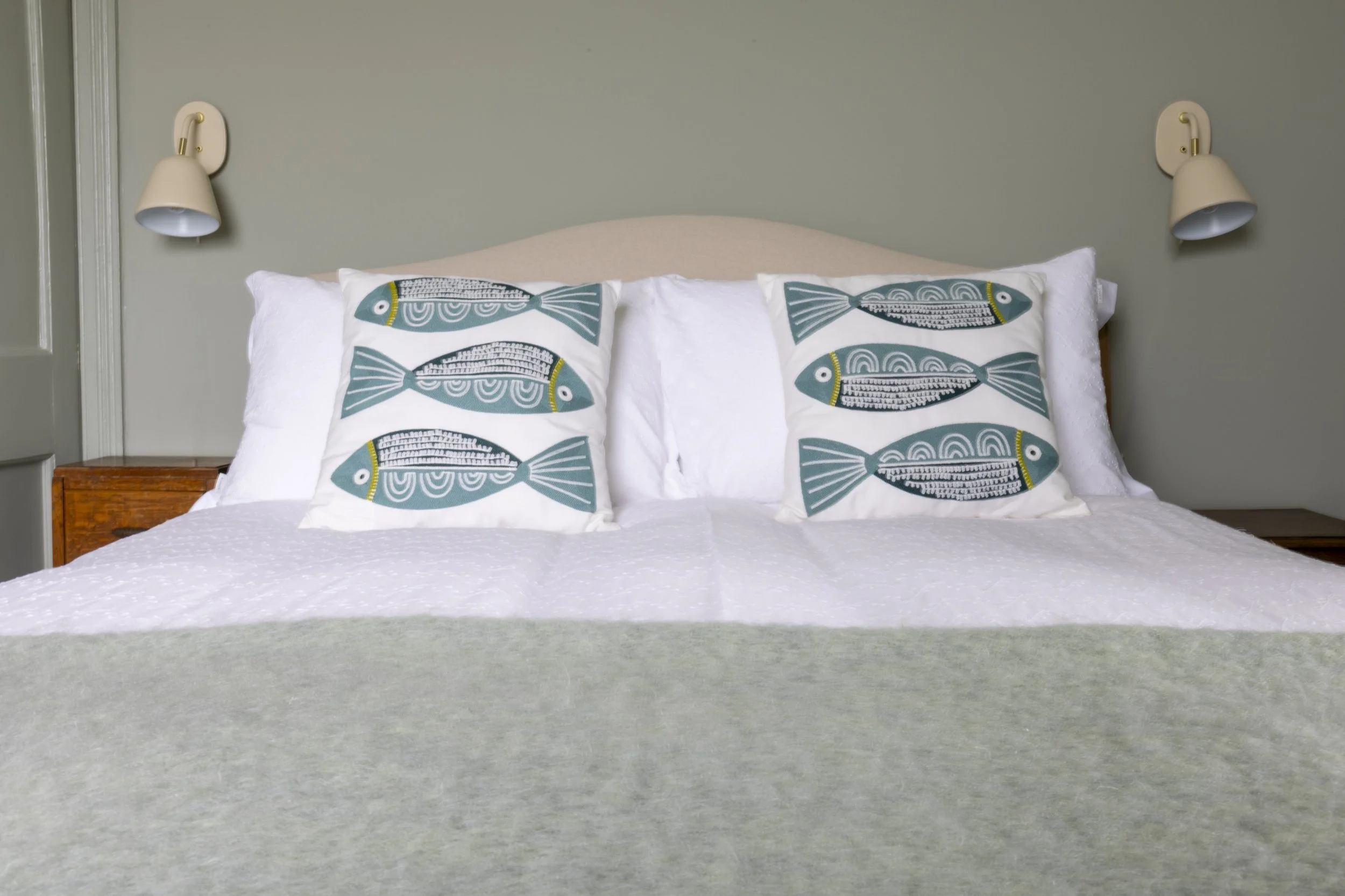 A neatly made bed with white sheets and two decorative pillows featuring a fish pattern, flanked by wall-mounted lamps on a green wall.